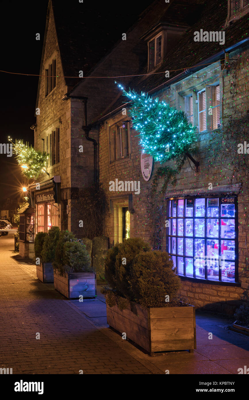 Bourton auf dem Wasser Geschäfte mit Weihnachtsbäumen und Lichter entlang der High Street in der Nacht. Bourton auf dem Wasser, Cotswolds, Gloucestershire, England Stockfoto