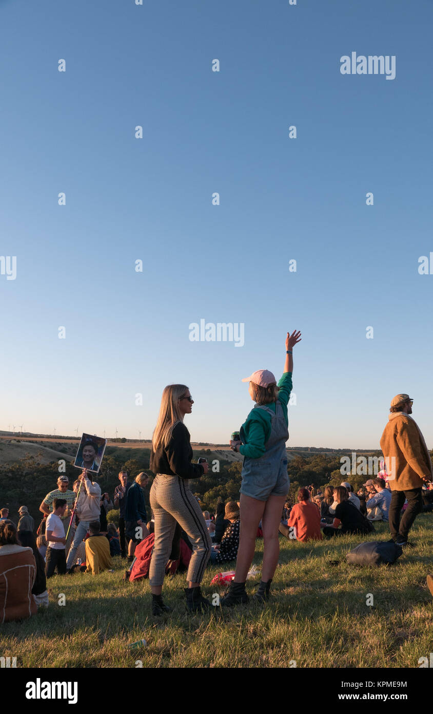 Große Gruppe junger, bunt gekleidet Music Festival goers beobachten einen magischen goldenen Sonnenuntergang am Horizont mit Windkraftanlagen im Hintergrund. Stockfoto