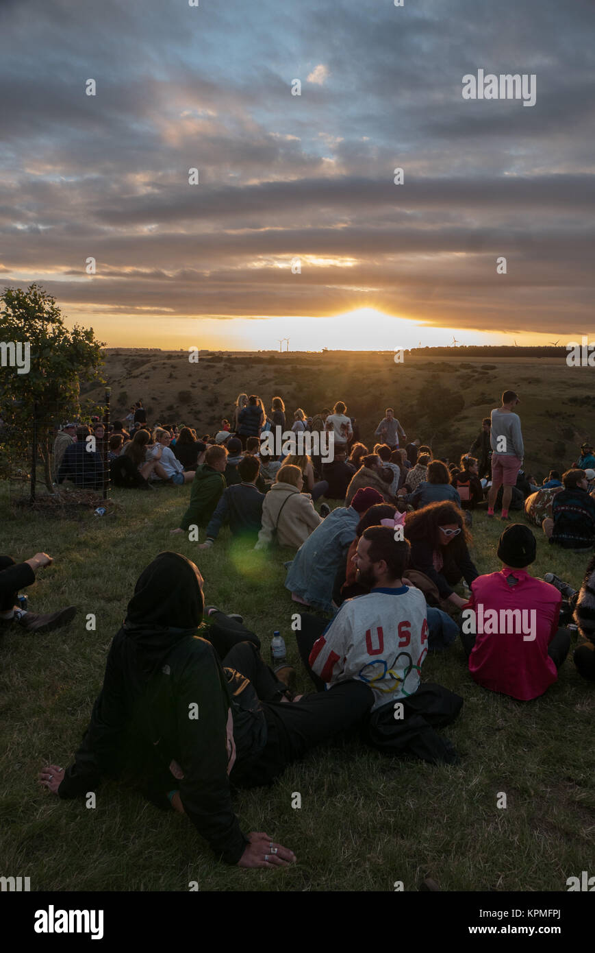 Große Gruppe junger, bunt gekleidet Music Festival goers beobachten einen magischen goldenen Sonnenuntergang am Horizont mit Windkraftanlagen im Hintergrund. Stockfoto