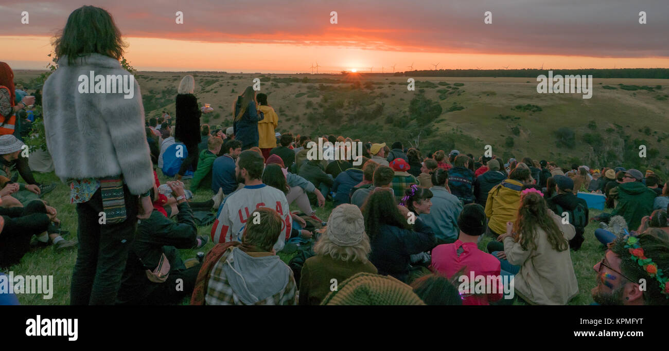 Große Gruppe junger, bunt gekleidet Music Festival goers beobachten einen magischen goldenen Sonnenuntergang am Horizont mit Windkraftanlagen im Hintergrund. Stockfoto