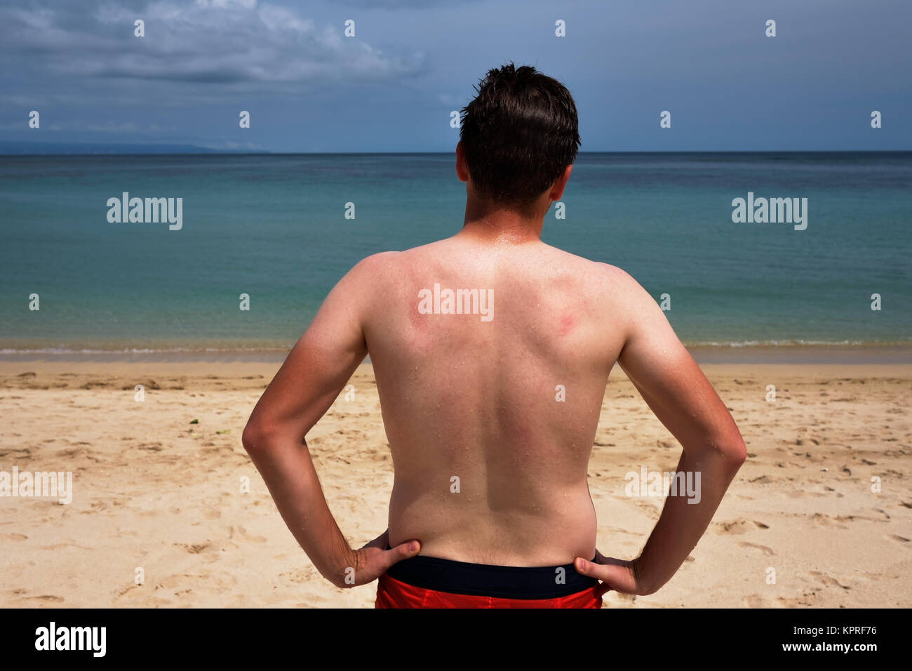 Ein junger Mann mit einem lustigen Tan aus einem T-Shirt steht auf dem Meer und blickt auf das klare, blaue Meer. Stockfoto