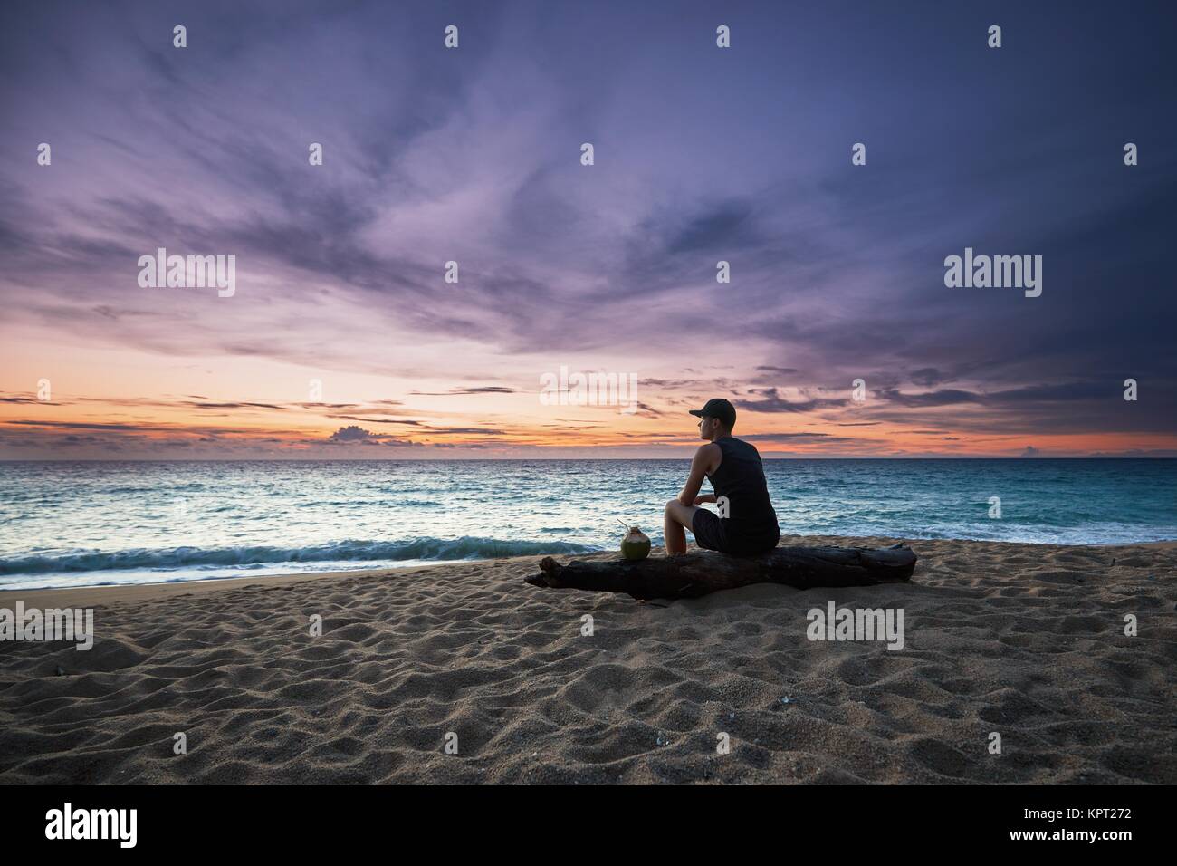 Nachdenklicher junger Mann mit Kokosnuss Drink relaxen am Strand bei Sonnenuntergang. Stockfoto
