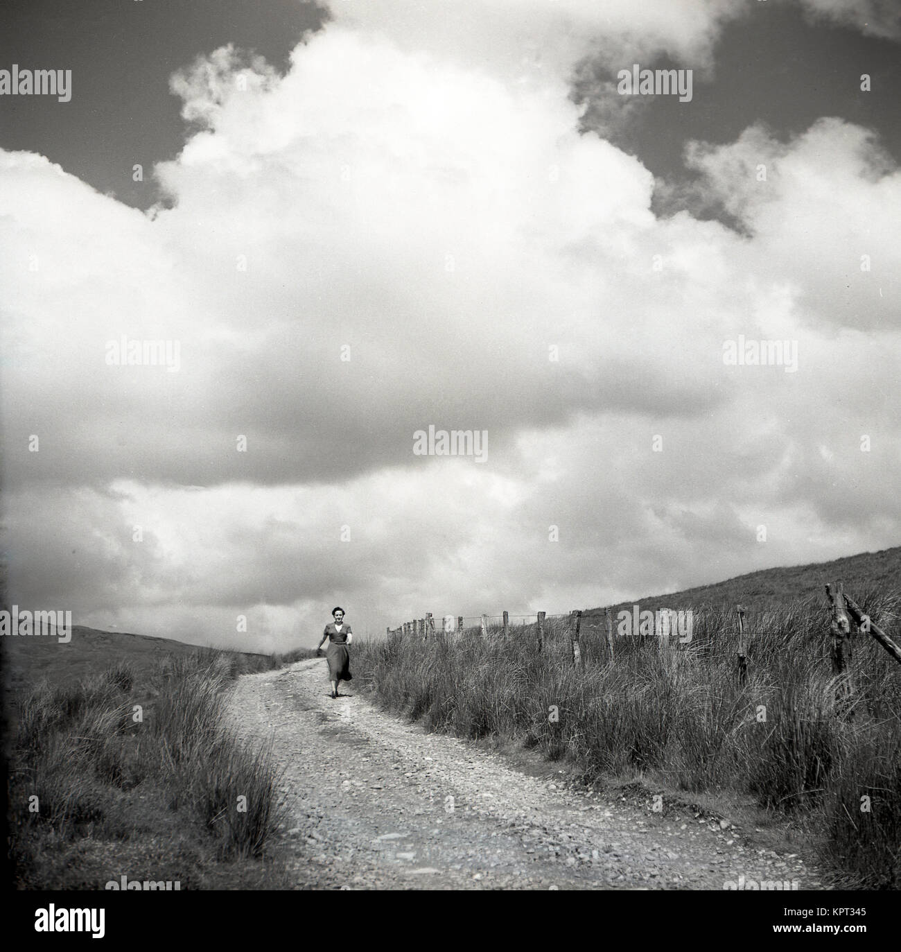 1950, historische, Sommer und eine Dame trägt ein Kleid ist außerhalb auf einem Kiesweg neben einigen Land, England, UK. Stockfoto