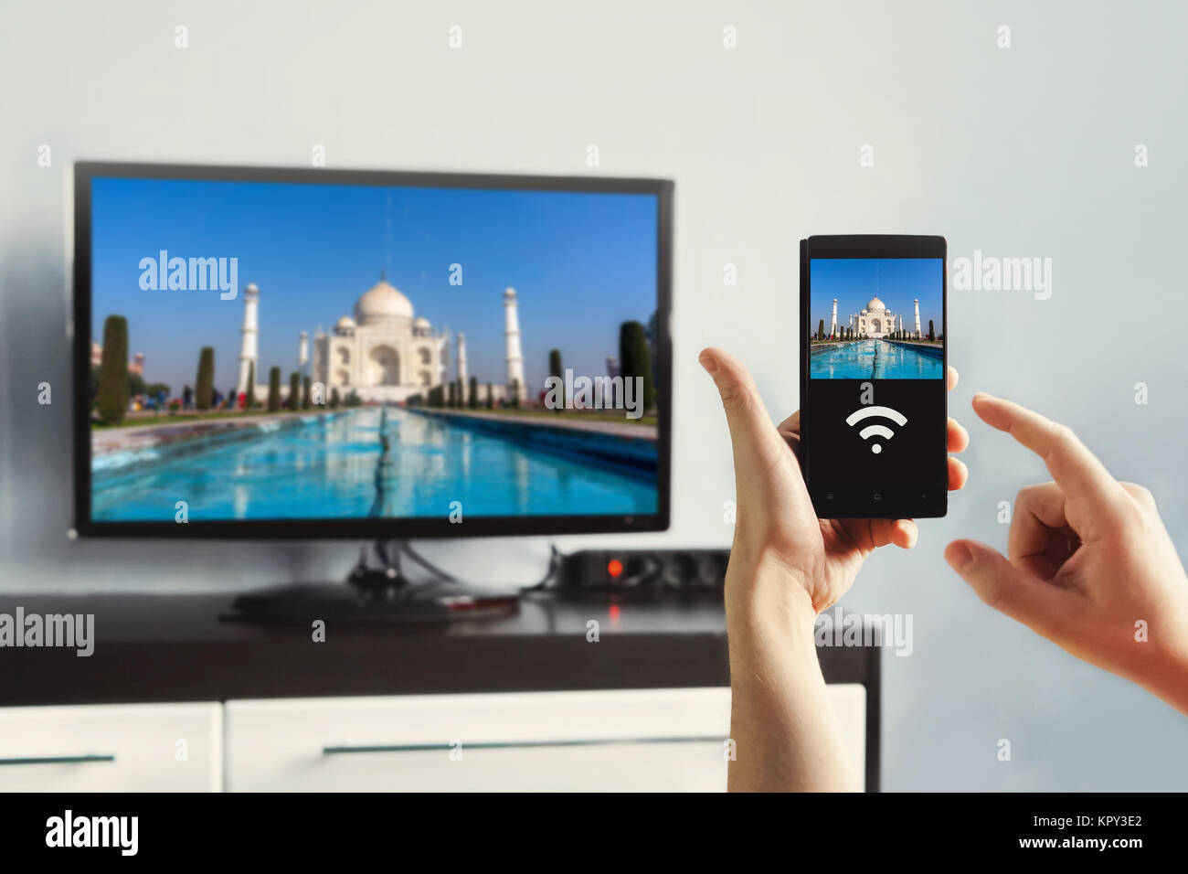 Männliche hand mit einem Smartphone gegen Aussicht von Wohnzimmer mit einem großen TV. Verwenden Sie Wi-Fi-Bilder vom Telefon auf den Bildschirm zu übertragen. Anzeigen Stockfoto