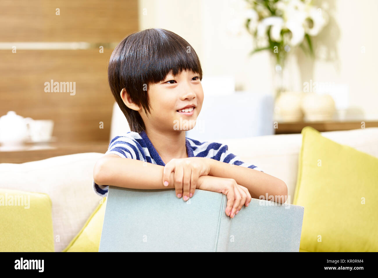7 Jahre alte asiatische kleiner Junge sitzt auf einem Sofa, die ein Buch lächelnd. Stockfoto