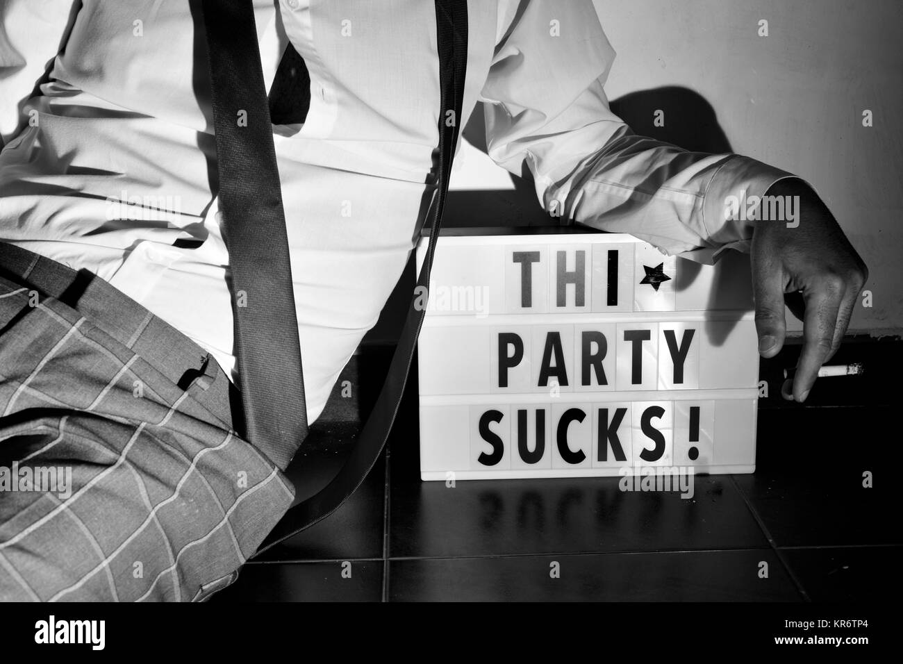 Nahaufnahme eines jungen Mannes auf dem Boden liegend tragen eine lose Black Tie und rauchen eine Zigarette, und der Text dieser Partei saugt in einer Lightbox neben ihm, Stockfoto