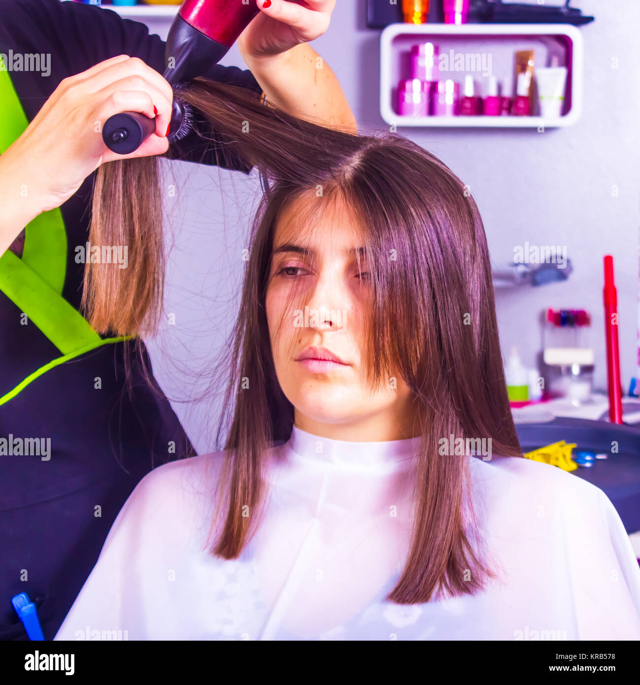 Stylist trocknen und stylen Frau Haar bedeckt mit einem Friseursalon Cape und sitzen in einem Salon Stuhl. Der Stylist verwendet eine hand Trockner und eine Haarbürste. Stockfoto