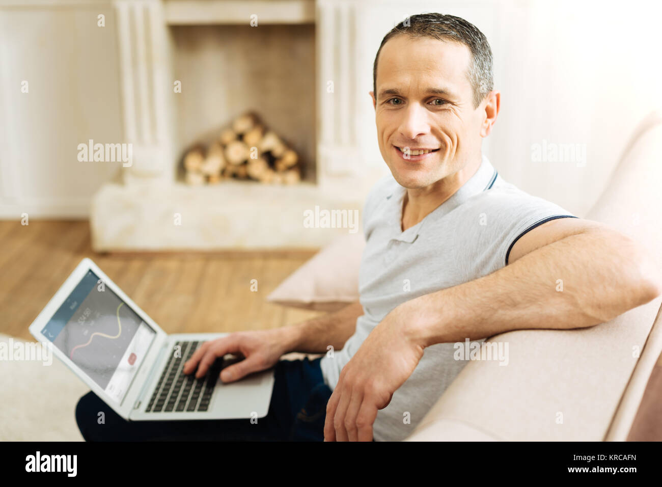Attraktive positiver Mann sitzend mit seinem Laptop und nachdenklich lächelnd Stockfoto