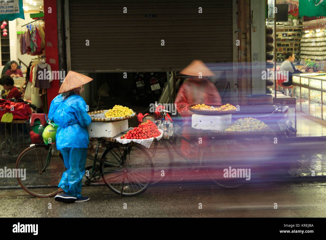Motorrad blur Verdecken bike Obst Verkäufer in Regen ponchos diskutieren Strategie auf einen feuchten Tag in den Straßen von Hanoi, Vietnam Stockfoto