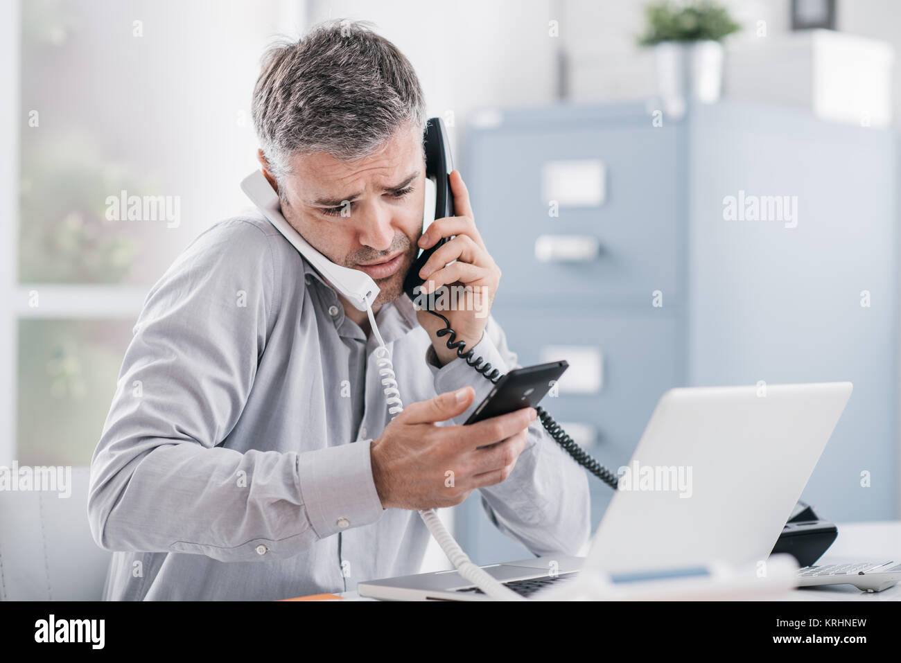Verzweifelt Geschäftsmanns, der in seinem Büro und in mehrere Anrufe betonte, er hält zwei Mobilteile und ein Handy, Business Management con Stockfoto