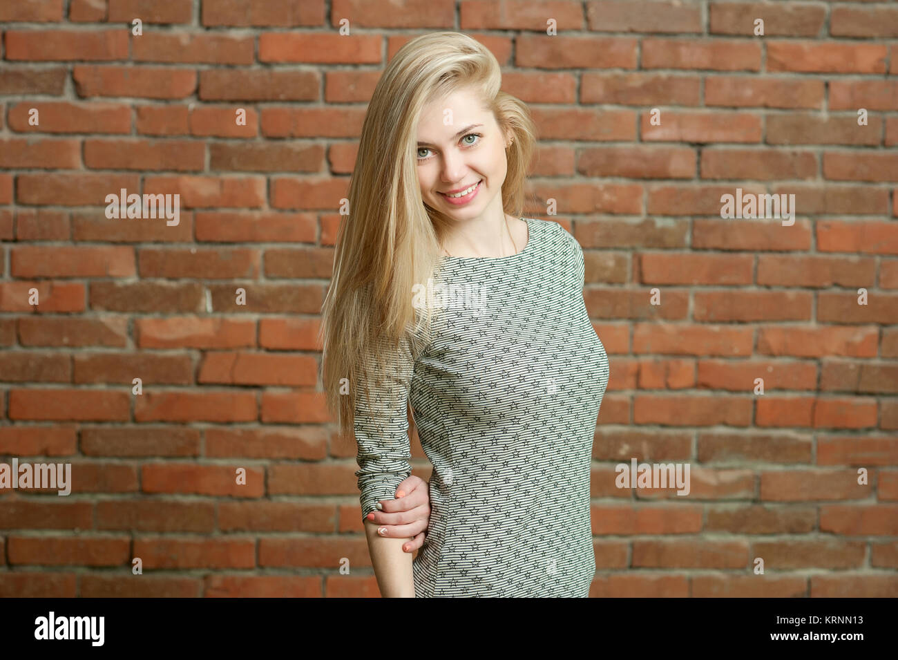 Porträt einer Blondine in einen leichten Pullover. Gegen die Red brick wall Hintergrund stehend, lächelnd und für die Kamera posieren. ihr langes Haar ist in einer Richtung festgelegt Stockfoto