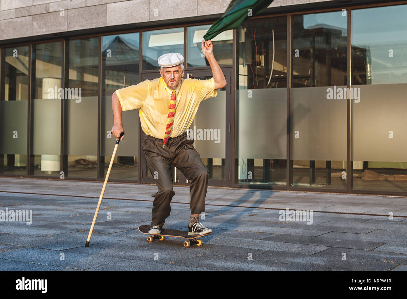 Verspielte Großvater rollt auf einem Skateboard, in der Hand eine Jacke, mit dem er winkte. Stockfoto