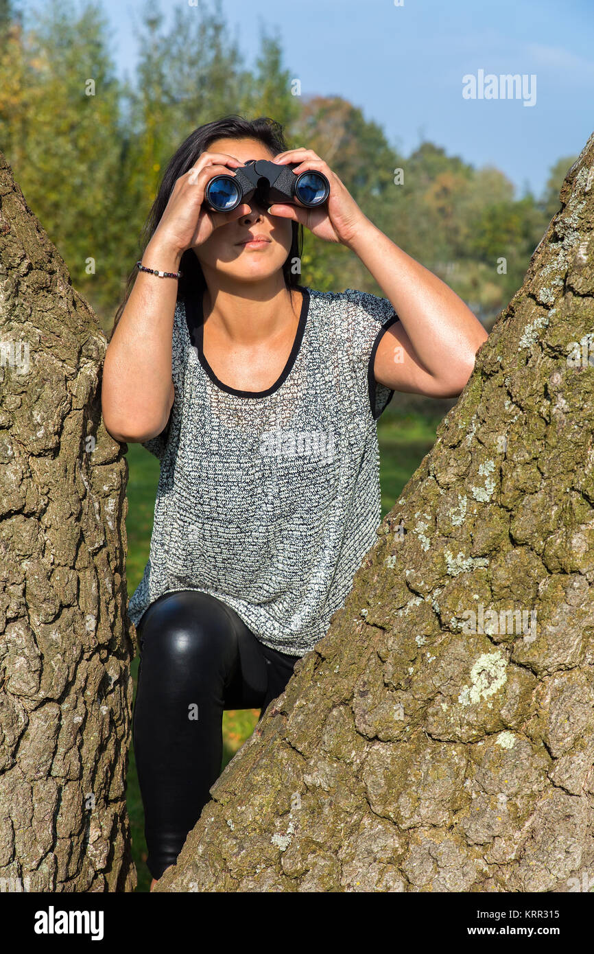 Junge Frau auf der Suche durchs Fernglas zwischen Baumstämmen Stockfoto