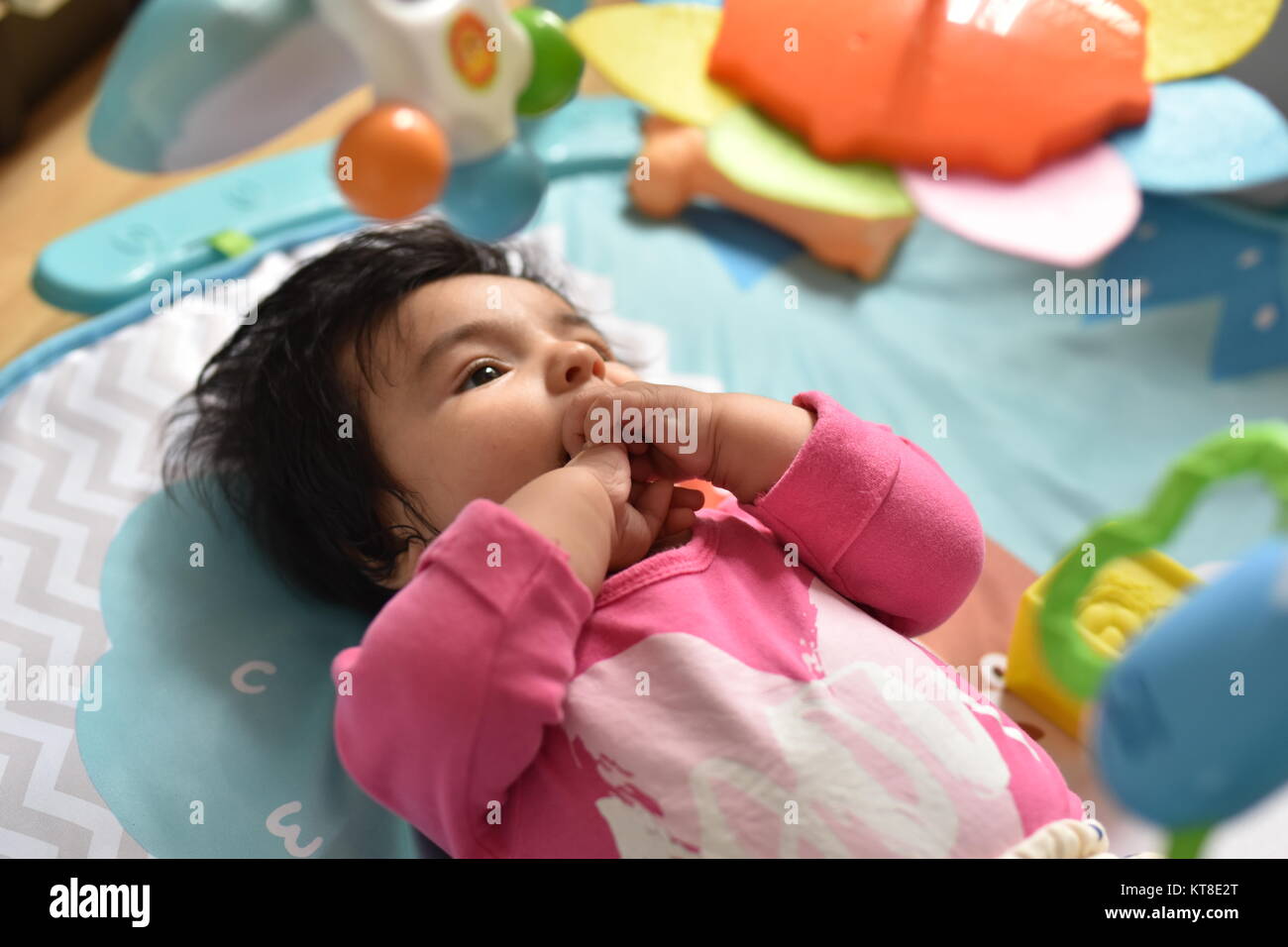 Baby Mädchen mit den Händen auf den Mund beobachtet aufmerksam ihre farbenfrohen Spielzeuge aus einem spielteppich Stockfoto