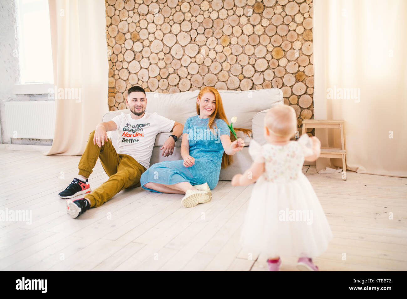 Junge Familie sitzt auf dem Boden in der Nähe der Couch, kleine Tochter einer Blondine ein Jahr alt ist, lernen in einem weißen Kleid vor dem Hintergrund von h zu Fuß Stockfoto