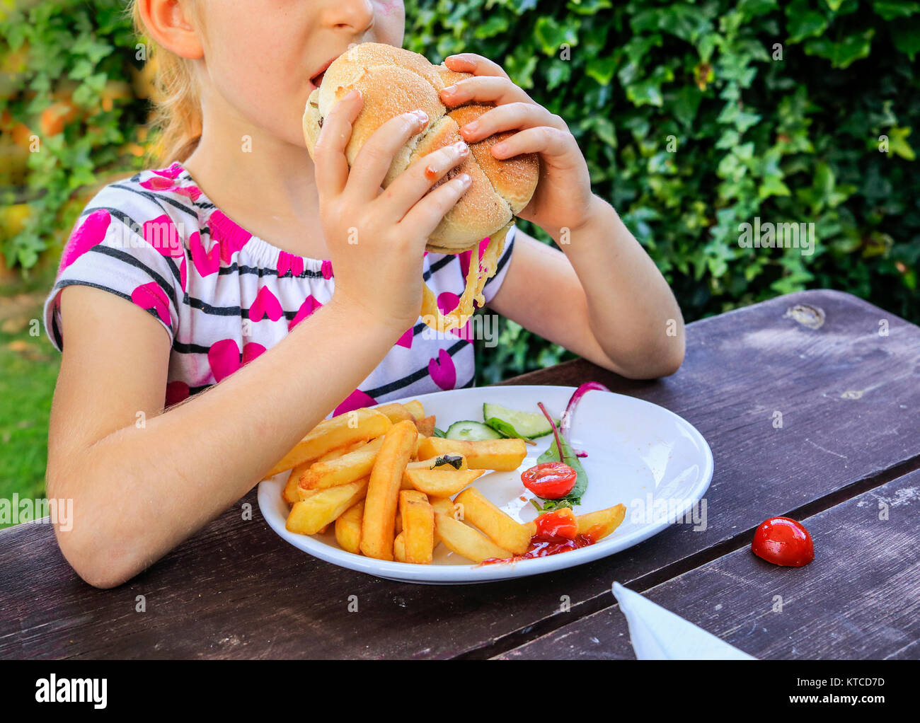 Junge Mädchen mit einer großen Hamburger in einem Restaurant im Freien; Platte mit Pommes Frites und Tomaten sitzt auf dem hölzernen Tisch; grüne Büsche in der b Stockfoto