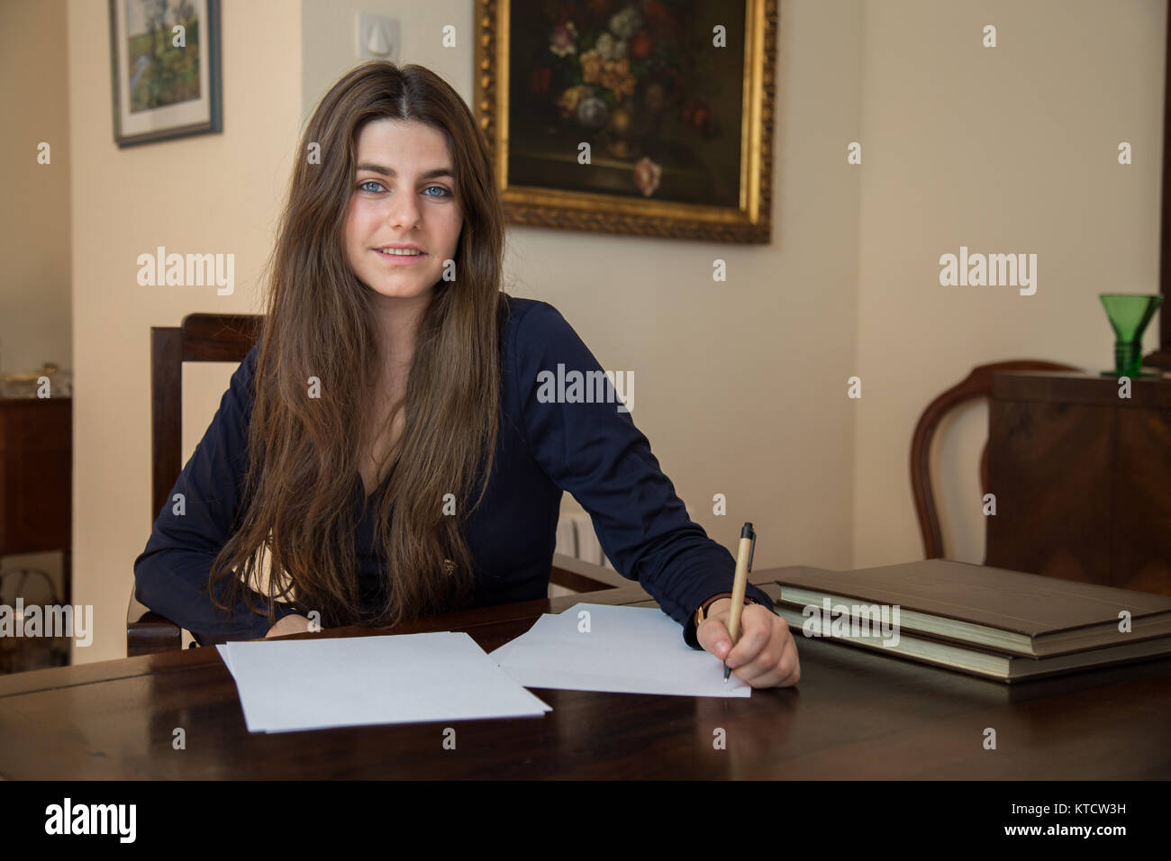 Hübsche, junge Frau mit blauen Augen und braune Haare sitzen auf einem eleganten hölzernen Tisch, indem sie auf ein leeres Blatt im Wohnzimmer ihres Hauses. Stockfoto