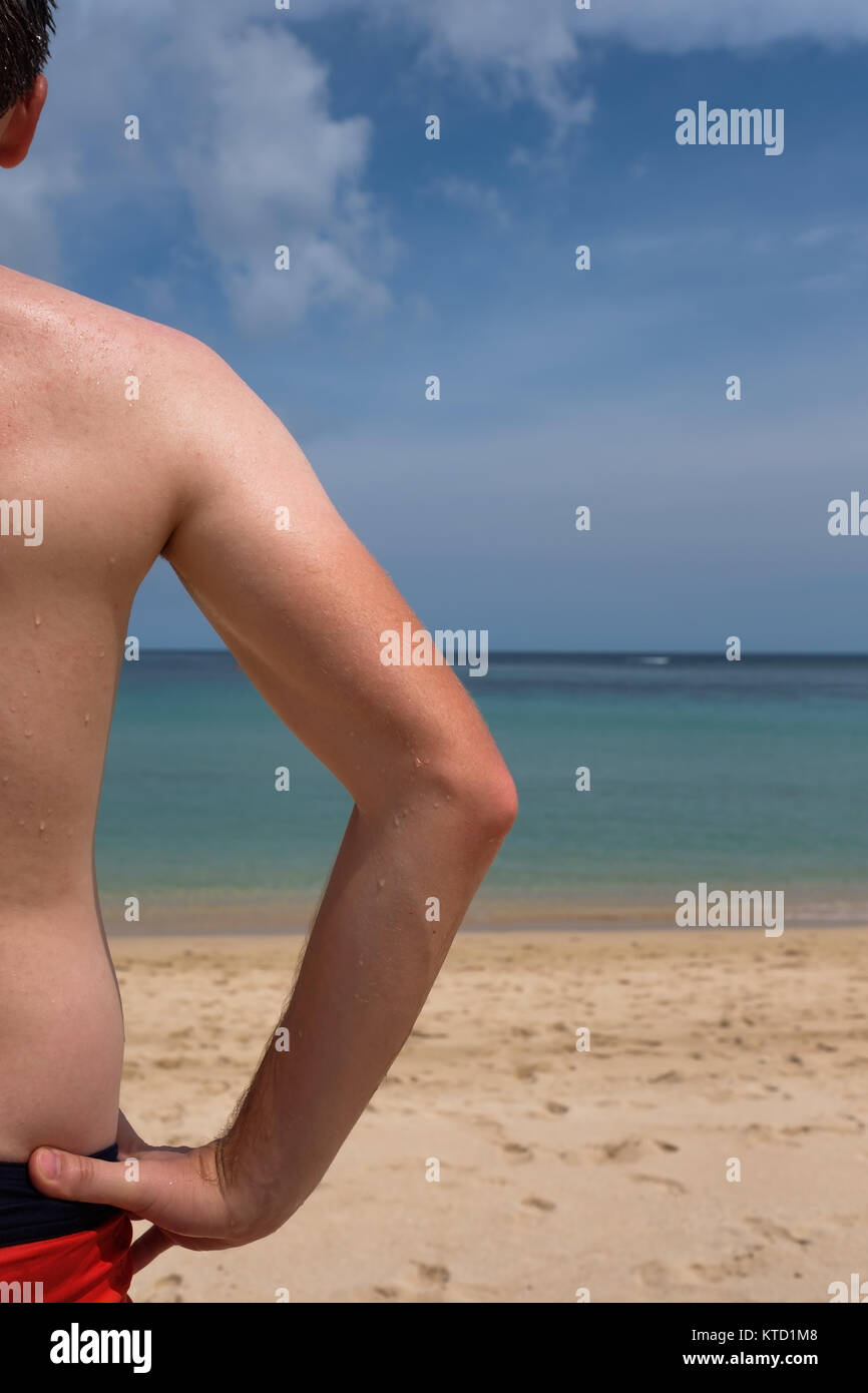 Ein junger Mann mit einem lustigen Tan aus einem T-Shirt steht auf dem Meer und blickt auf das klare, blaue Meer. Konzept der guten Urlaub Stockfoto