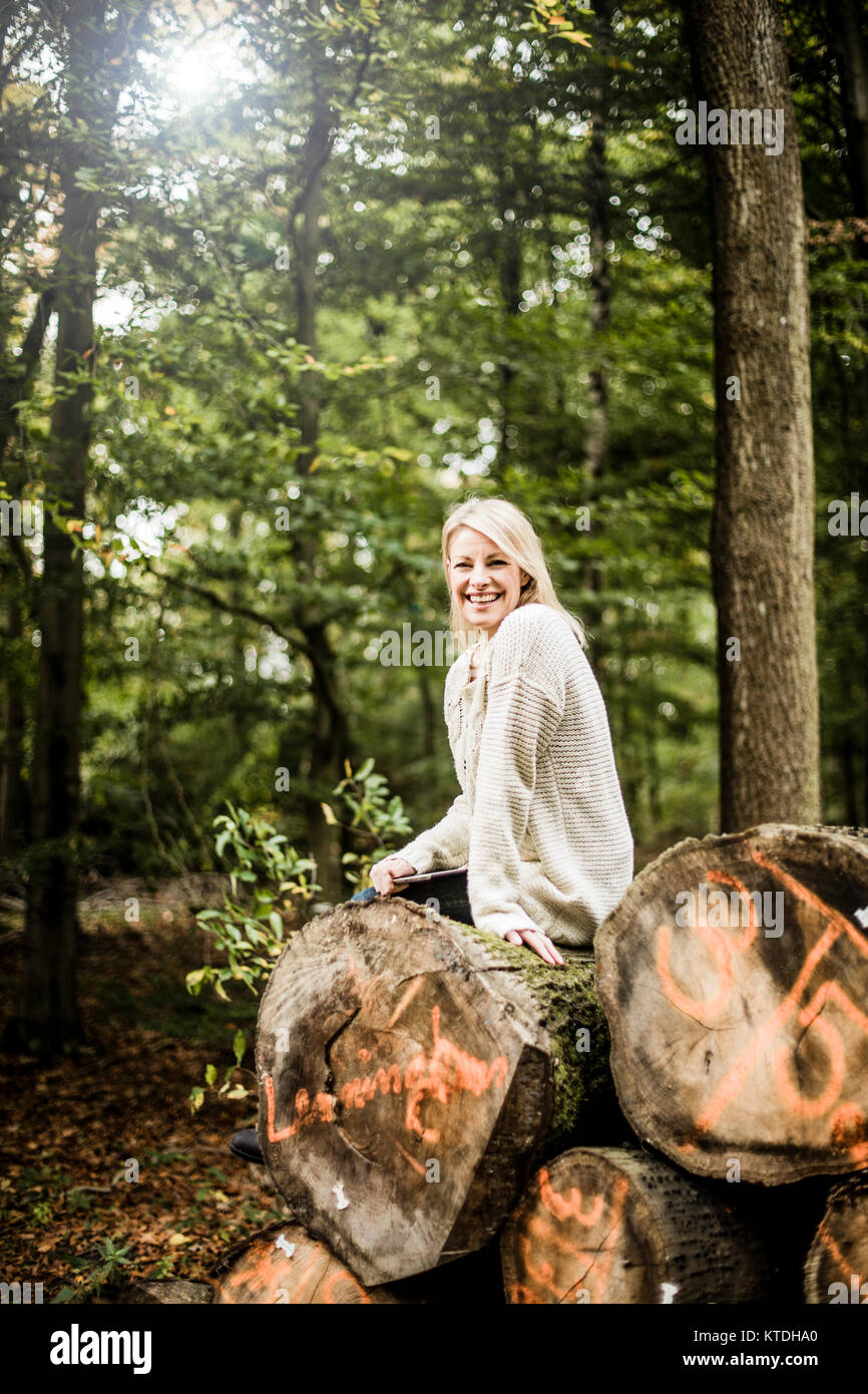 Portrait von glücklichen Frau sitzt auf Stapel Baumstämme im Wald mit Tablet Stockfoto