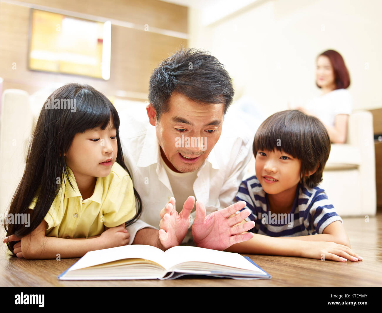 Asiatische Vater und zwei Kinder liegen auf der Vorderseite auf dem Boden lesen Buch, während Mutter aufpassen im Hintergrund. Stockfoto