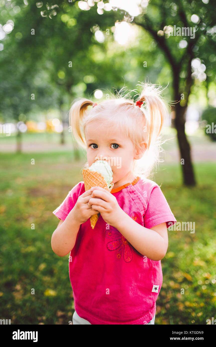 Wenig lustig Mädchen blond Süßigkeiten blaue Eis in der Waffel Schale auf einem grünen Hintergrund im Park. verschmierte Gesicht und Wangen und lacht. Stockfoto
