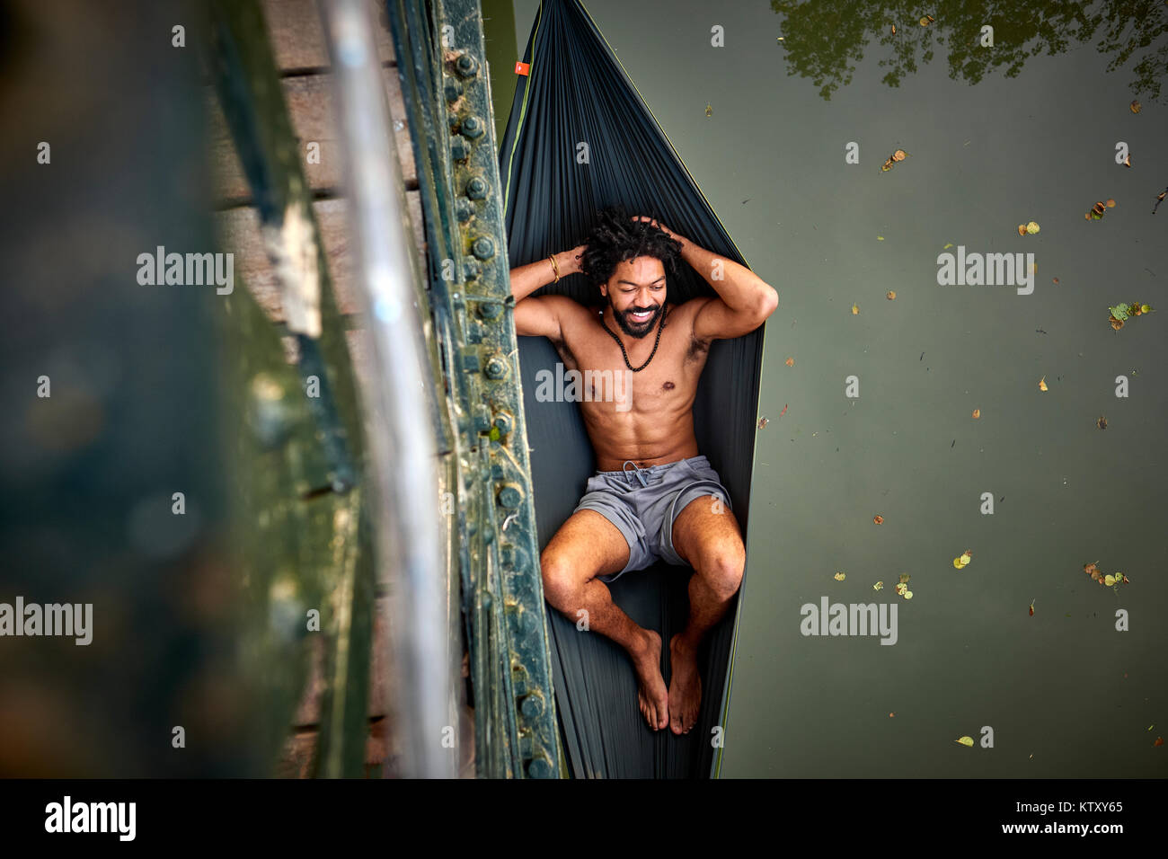 Ein Mann entspannt in der Hängematte umschlungen von einer Brücke über einen Fluss. Stockfoto