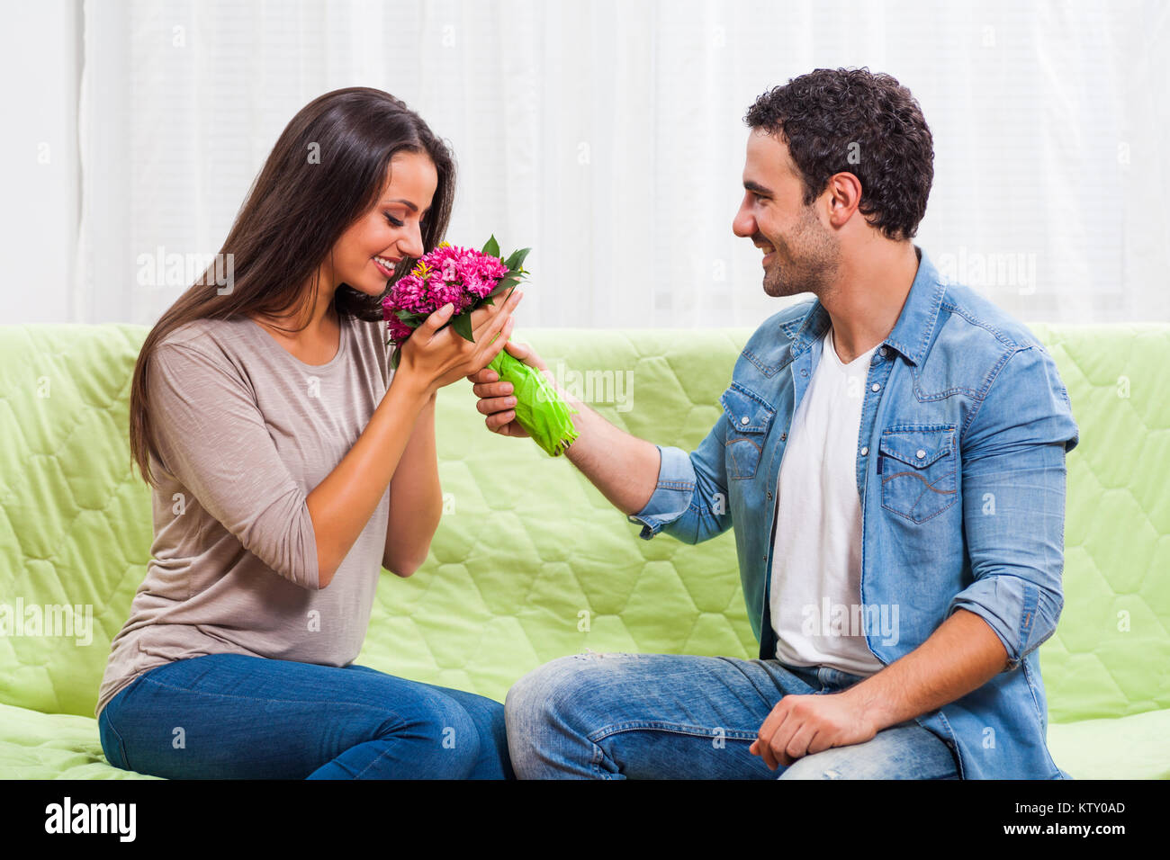 Junges Paar sitzt auf einem Sofa zu Hause. Der Mensch ist mit Blumen zu seiner Frau. Stockfoto