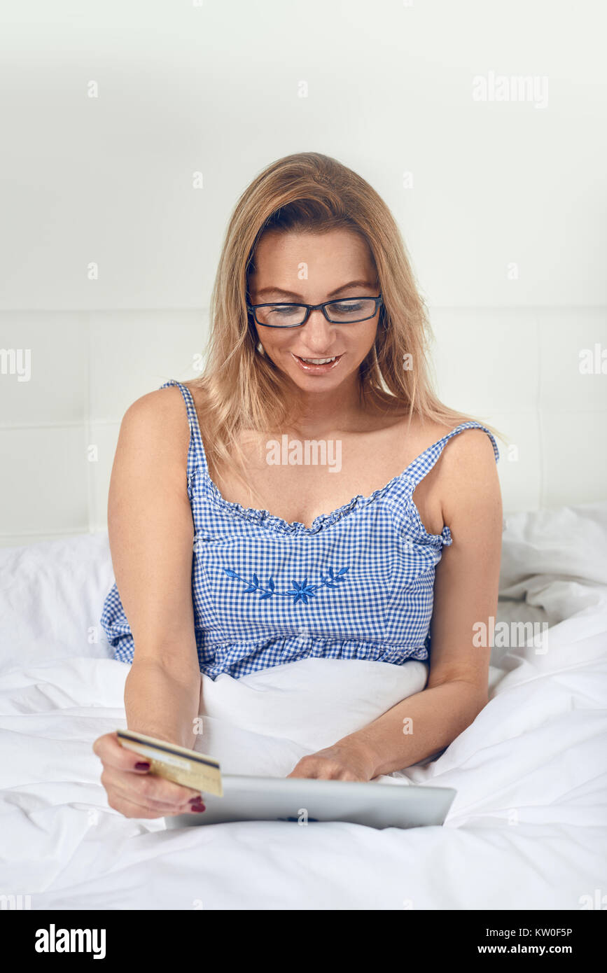 Attraktive Frau entspannen im Bett tun, online shopping hält Ihre Kreditkarte in einer Hand und digitale Tablet in den anderen, wie sie in die Kamera lächelt Stockfoto