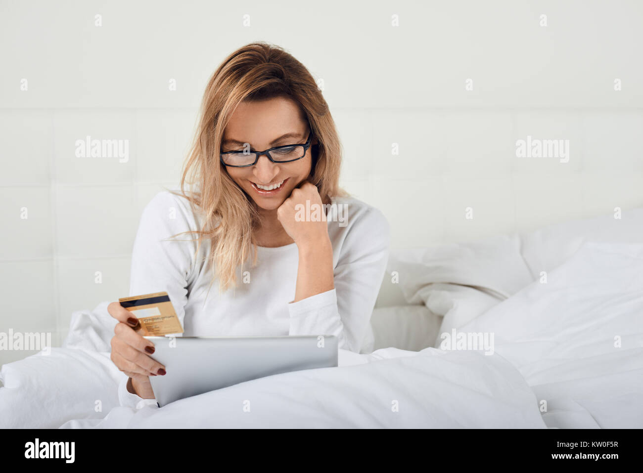 Attraktive Frau entspannen im Bett tun, online shopping hält Ihre Kreditkarte in einer Hand und digitale Tablet in den anderen, wie sie in die Kamera lächelt Stockfoto