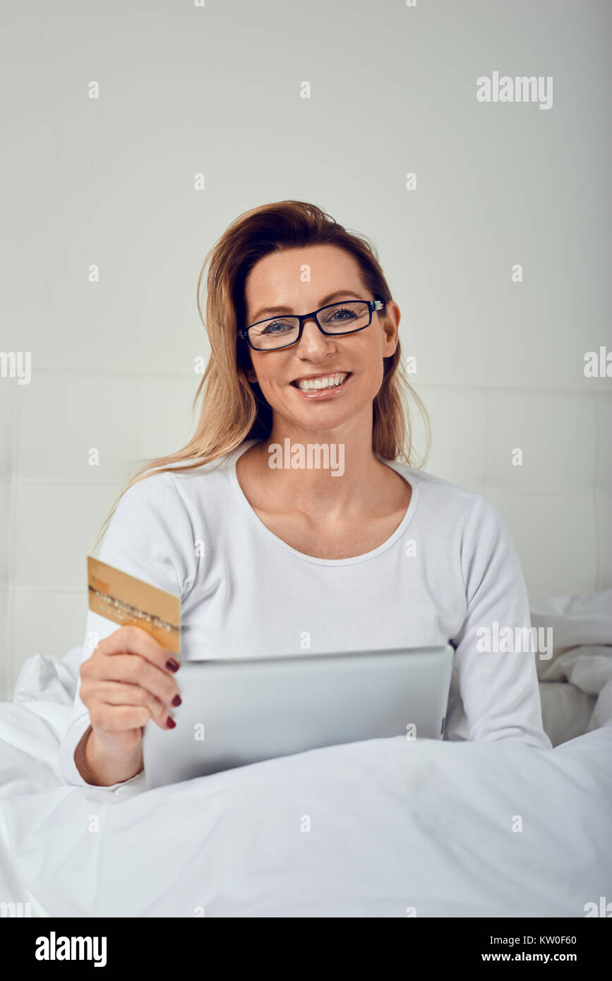 Attraktive Frau entspannen im Bett tun, online shopping hält Ihre Kreditkarte in einer Hand und digitale Tablet in den anderen, wie sie in die Kamera lächelt Stockfoto