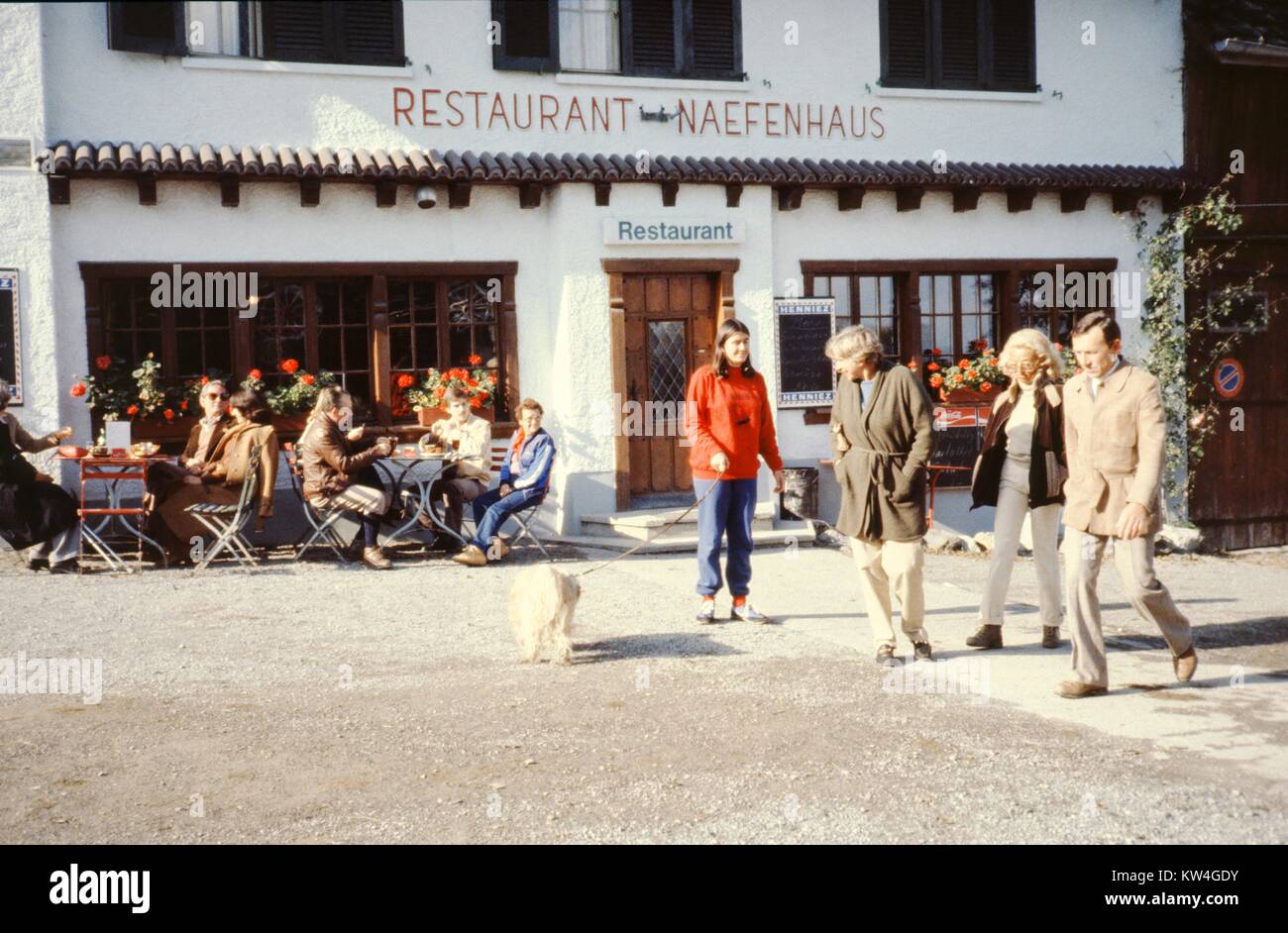 Eine Frau trägt eine rote Jacke Spaziergänge mit ihrem Hund als Touristen  vorbei Diners außerhalb des Restaurants Naefenhaus in Stallikon, Schweiz,  1975 Essen Stockfotografie - Alamy