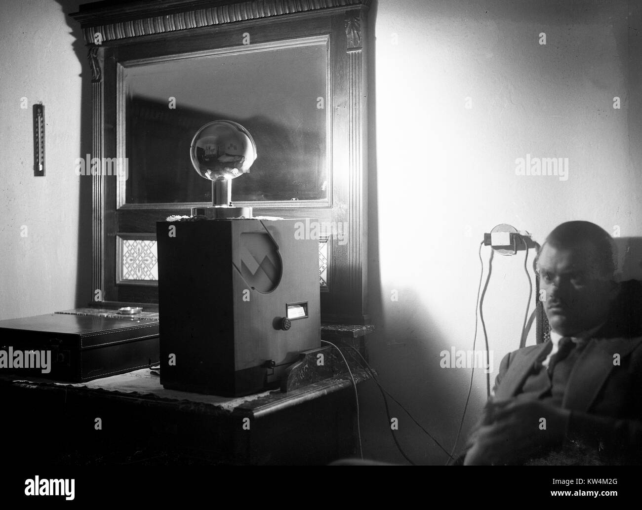 Ein Mann mit einem Stern Ausdruck sitzt in einem dunklen Raum und hört einen großen Radio, das auf einem Schreibtisch sitzt, als eine kleine Lampe über dem Radio auf eine Wand Spiegel reflektiert, die Beleuchtung des Raumes in krassem Schatten, mit einem Aktenkoffer auf dem Schreibtisch, Ungarn, 1929 sichtbar. Von einem Gelatine Prozess Glasplatte negative digitalisiert. Stockfoto