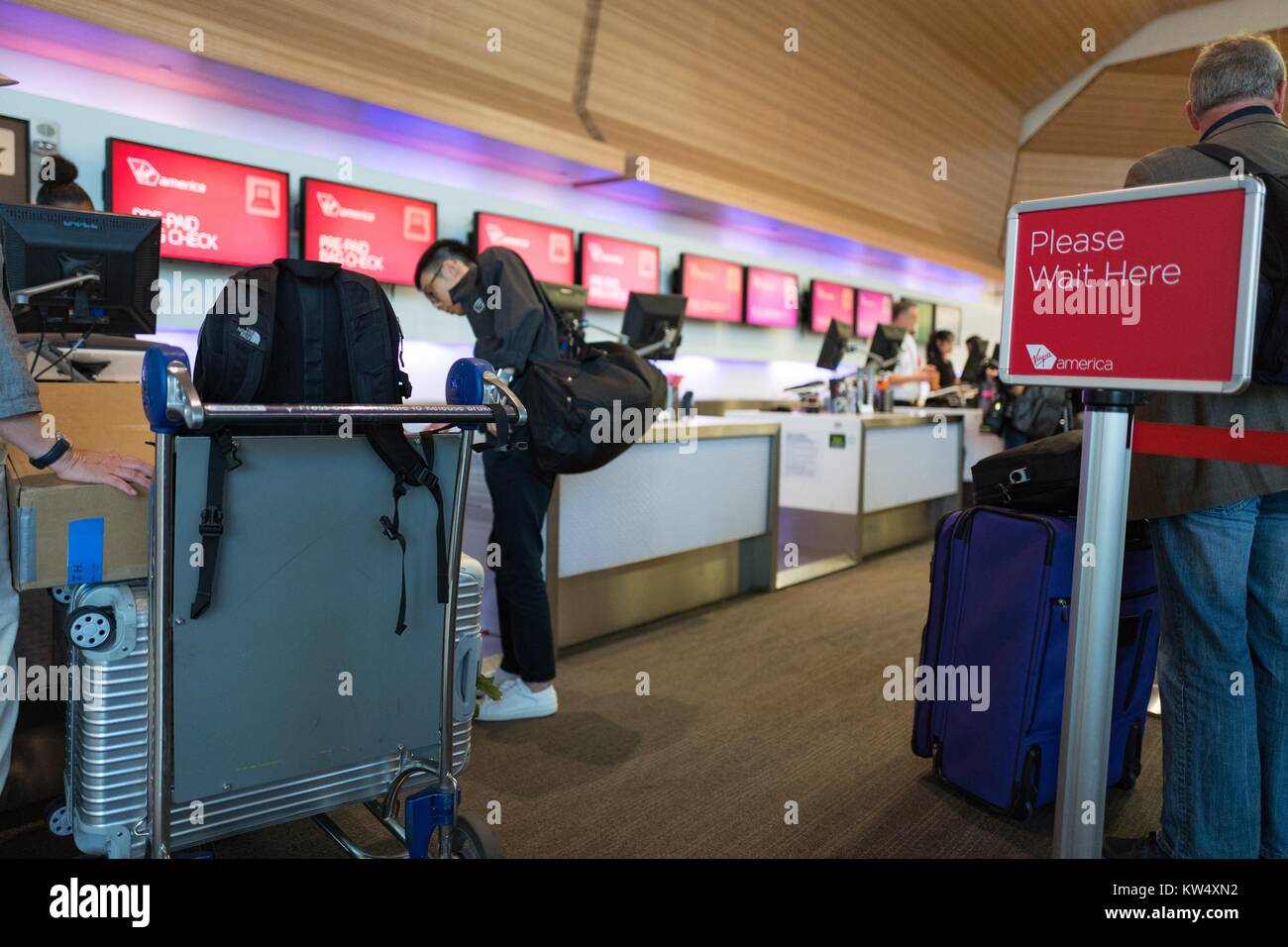 Passagiere mit einem Warenkorb Smarte Carte Gepäck in Ihre Flüge und Gepäck an einer Jungfrau, die Amerikanische Ticket Schalter prüfen, während andere Passagiere in Linie warten, am San Francisco International Airport, South San Francisco, Kalifornien, USA, 24. September 2016. Ein Zeichen im Vordergrund lautet: "Bitte warten Sie hier.". Stockfoto