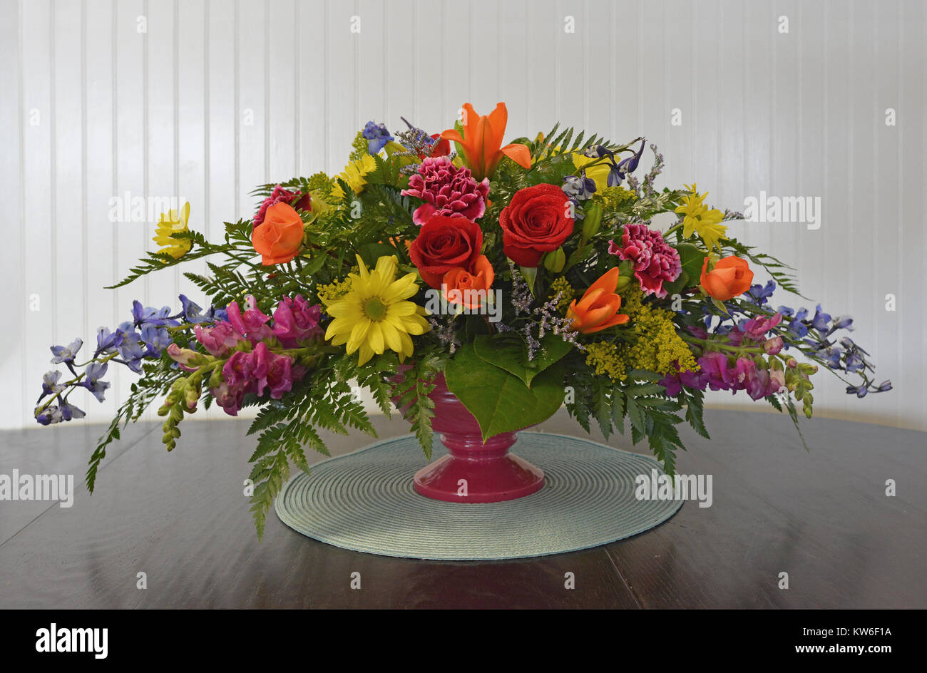 Eine bunte, horizontalen Mittelpunkt gefüllt mit Rosen, Lilien, Gänseblümchen, snapdragons, Rittersporn und Kaskadierung Laub auf einem Tisch gegen weiße Holz. Stockfoto