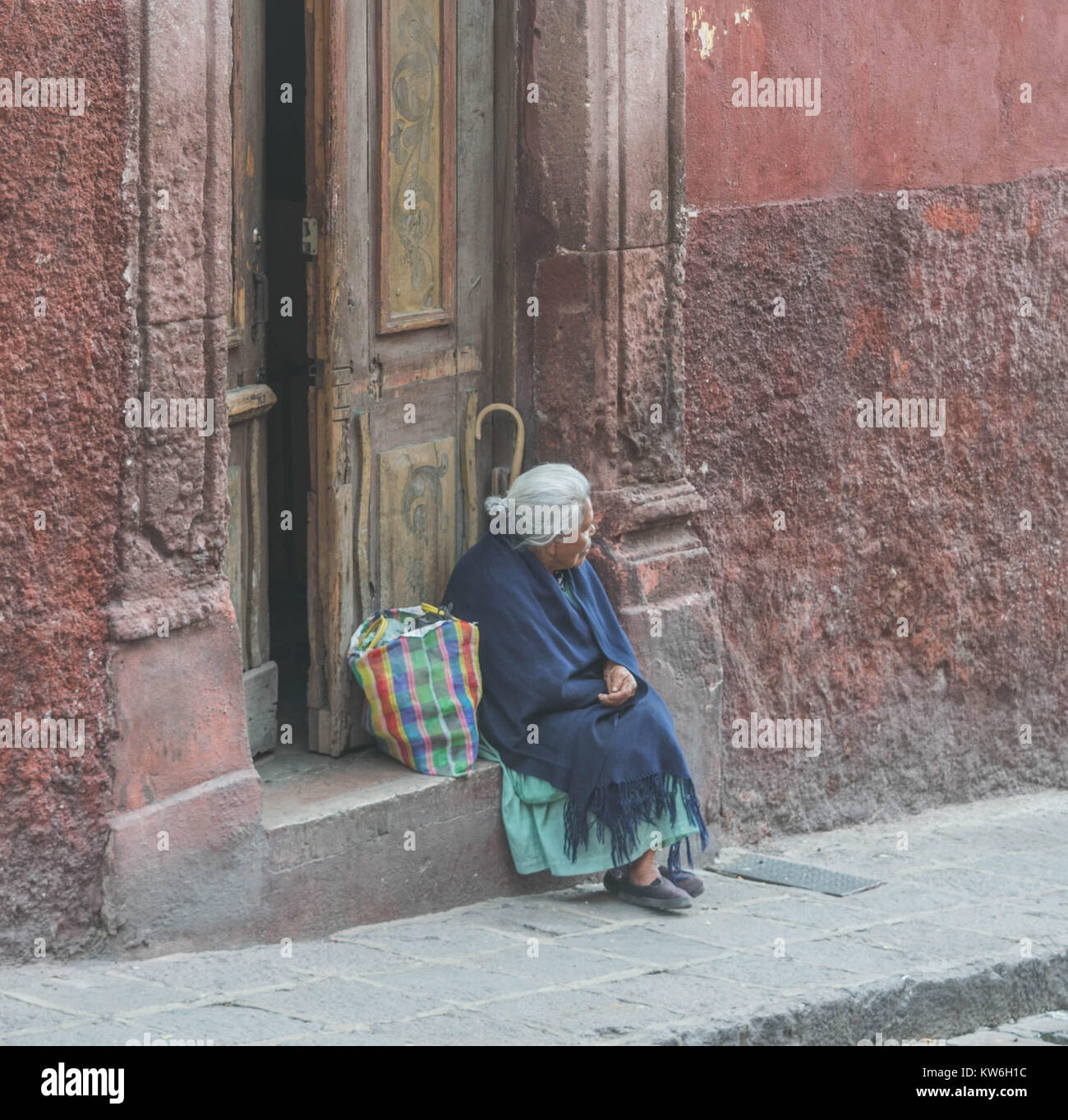 Eine ältere Frau mit grauem Haar, einen blauen Schal um die Schultern und einem bunten Netzbeutel sitzt auf einem Stein Schritt, in San Miguel de Allende, Mexiko Stockfoto