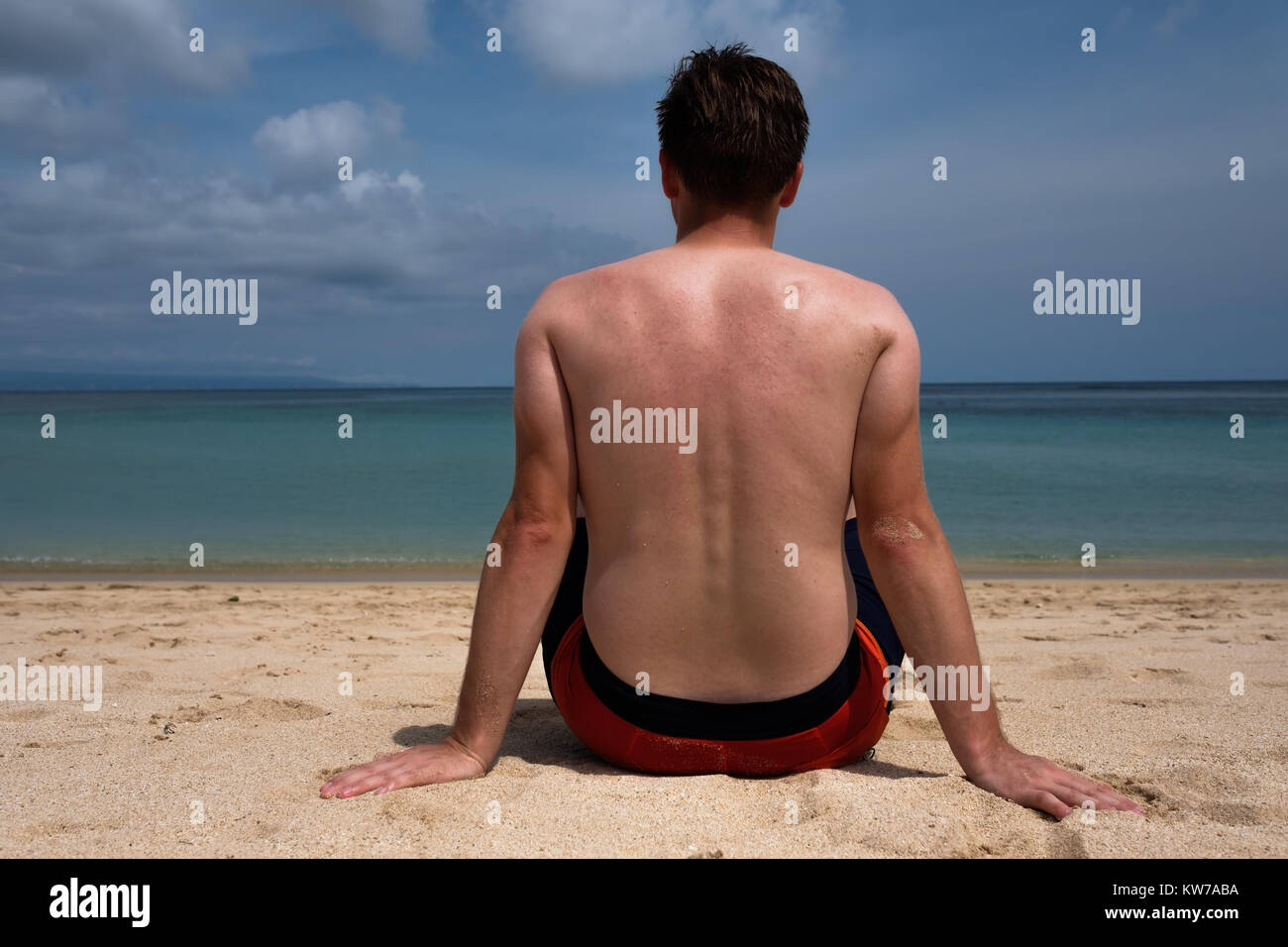 Ein junger Mann mit einem lustigen Tan aus einem T-Shirt steht auf dem Meer und blickt auf das klare, blaue Meer. Stockfoto