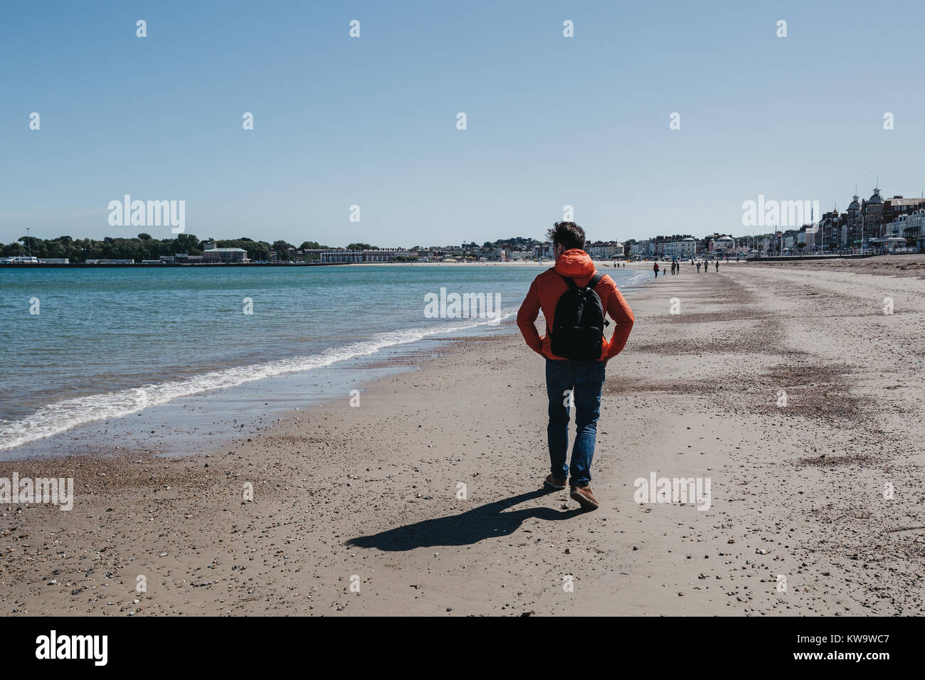 Junger Mann in rote Jacke und Jeans zu Fuß am Strand von Weymouth, Dorset, England an einem klaren sonnigen Tag. Stockfoto