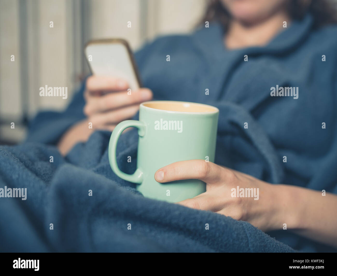 Eine junge Frau im Bademantel ist das Trinken einer Tasse Tee im Bett und Sie ihr Smartphone Stockfoto