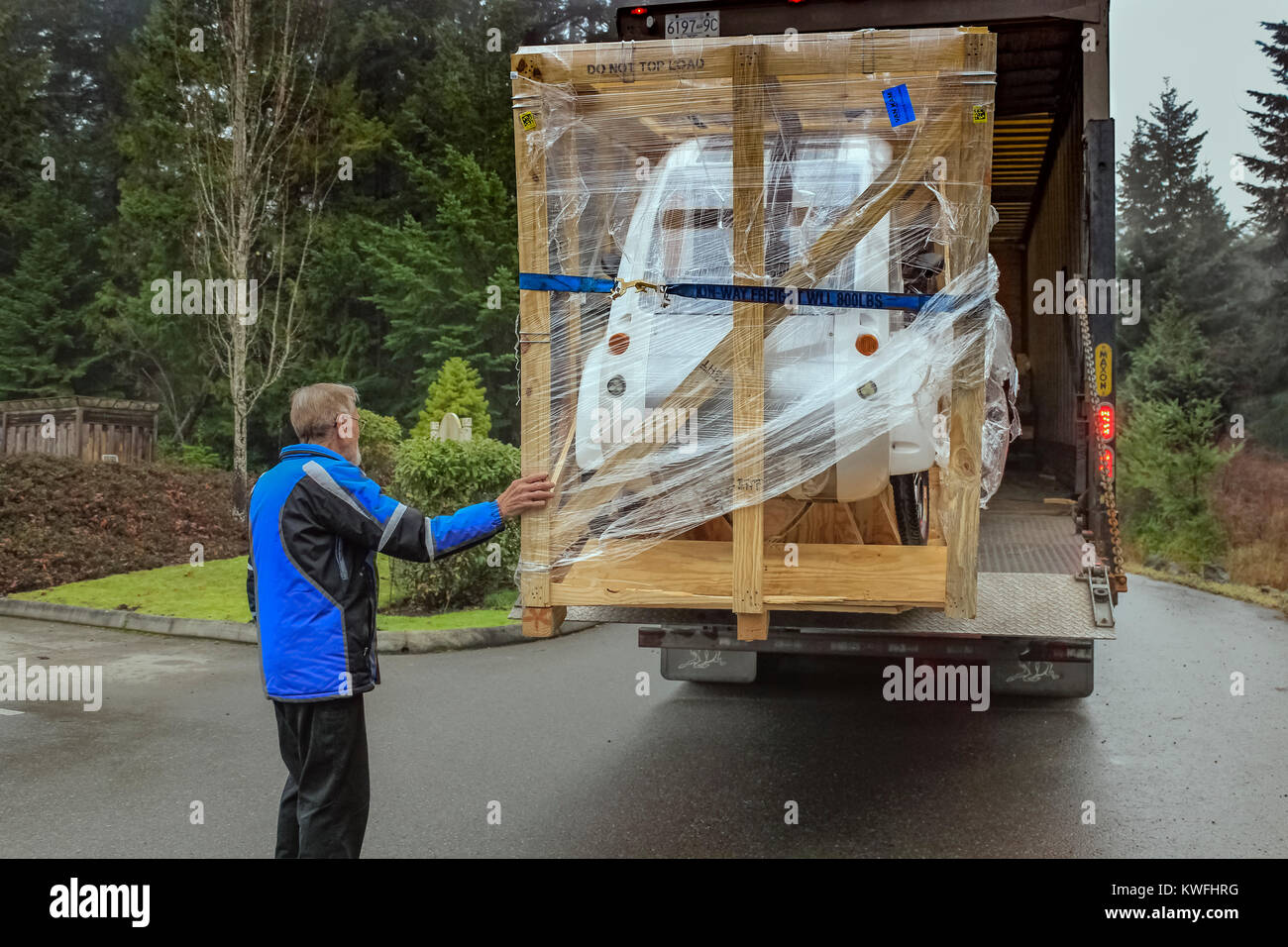 Ein älterer Mann in einen blauen Mantel Versand berührt eine große Kiste mit eine neue elektrische ELF Dreirad, als er für einen Transport LKW zu entladen wartet. Stockfoto