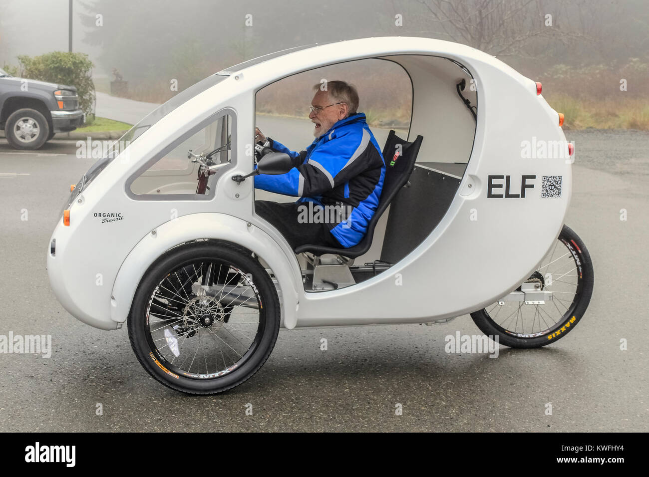 An einem nebligen Wintertag, ein älterer Mann (Alter 94) in einer blauen Jacke nimmt seine erste Fahrt in seinem neuen weißen Pedal - powered/elektrische ELF Dreirad. Stockfoto
