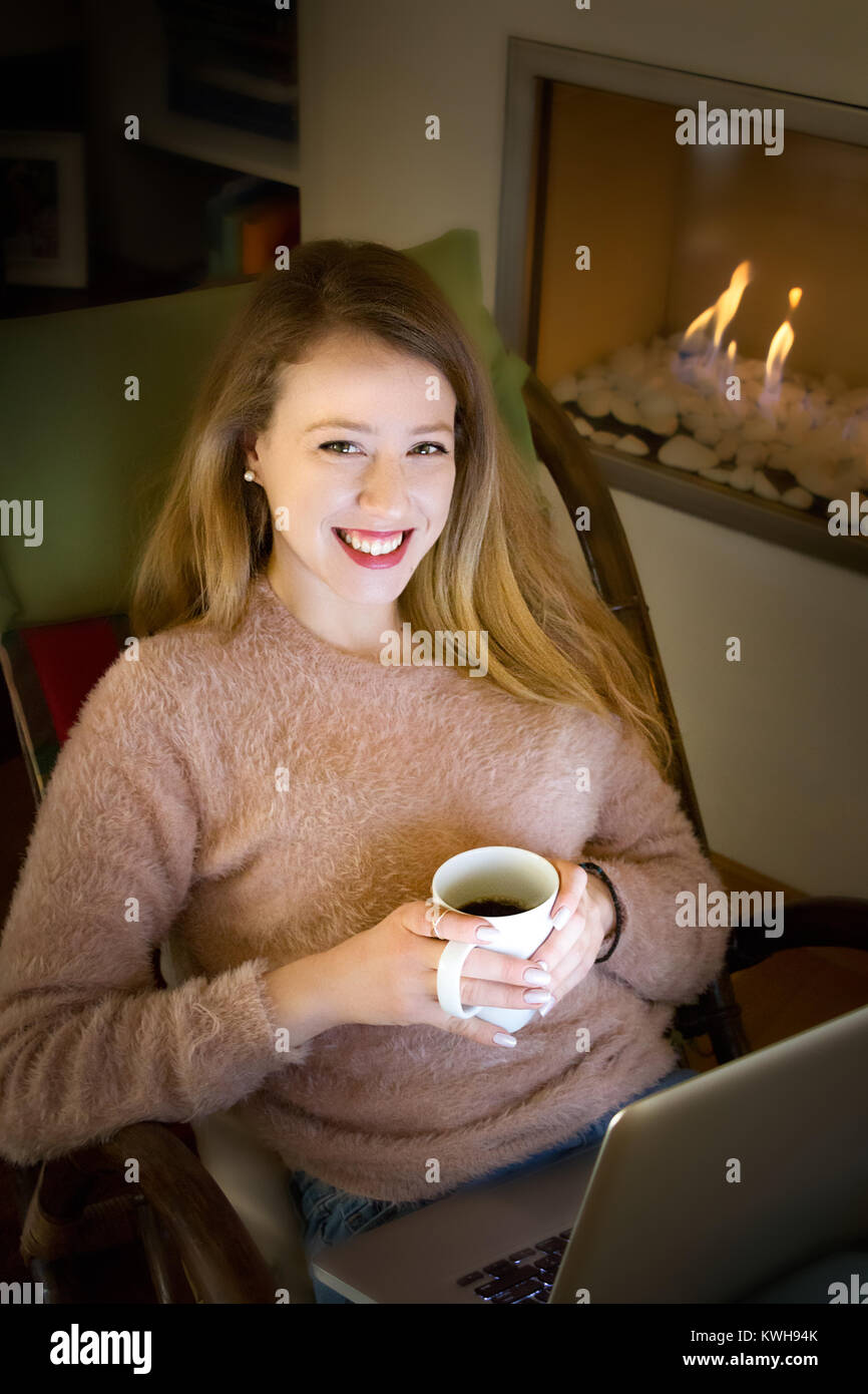 Porträt einer blonden jungen Frau an der Kamera lächelnd, auf einem Schaukelstuhl mit Ihrem Laptop sitzen und Kaffee an einem Kamin, Innen, gemütliche Konzept Stockfoto