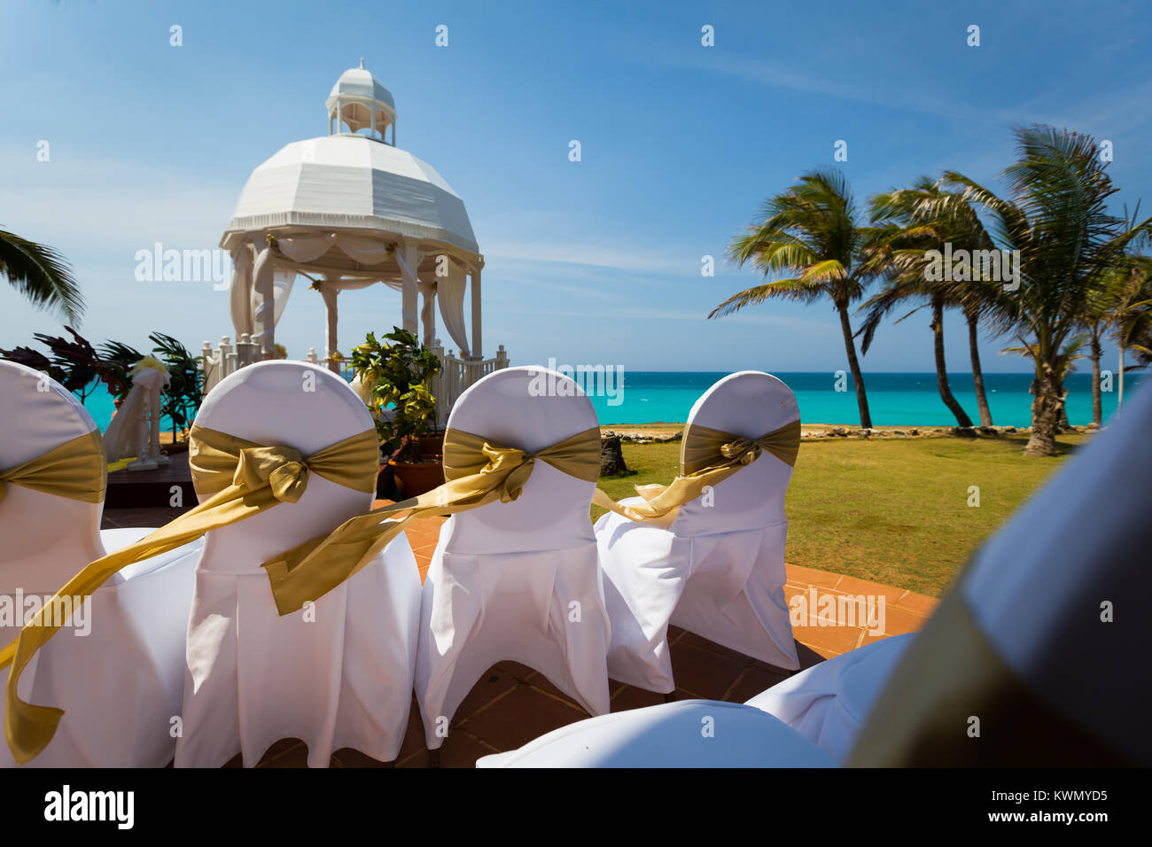 Outdoor tropische Hochzeit mit verzierten Stühle in weißen Stoff bedeckt und gebunden mit Gold Ribbon in einem windigen Garten mit Palmen mit Blick auf t Stockfoto