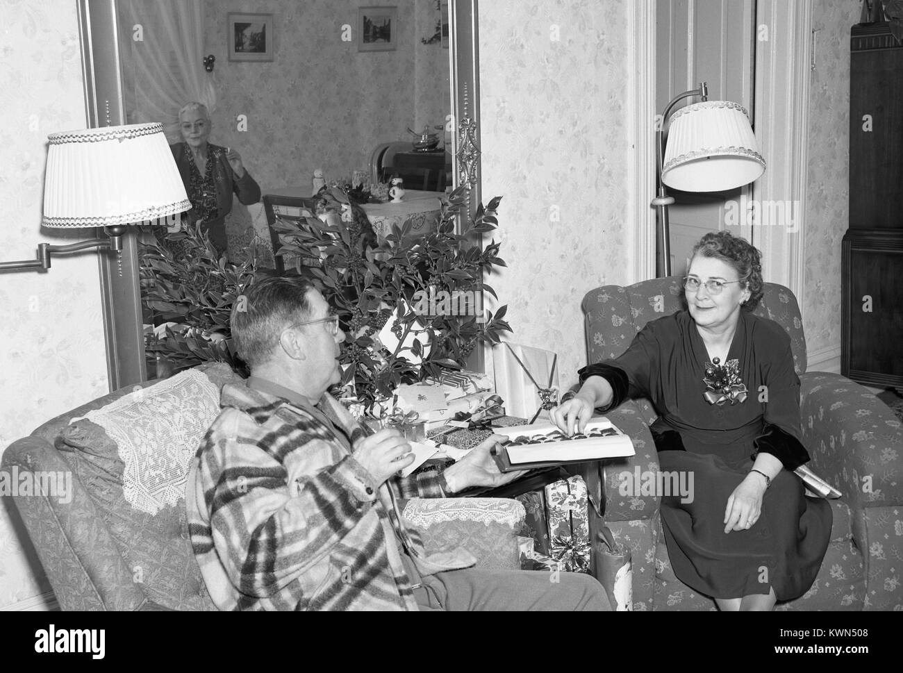 Einem erwachsenen Mann und große Paar sitzt im Sessel in einem vorstadthaus und eine Schachtel Pralinen teilen, der Ehemann übergeben die Pralinen auf die Ehefrau, während eine reife Frau, die in einem Spiegel an der Wand sichtbar, schaut an und raucht eine Zigarette, Eureka, Kalifornien, 1950. Stockfoto