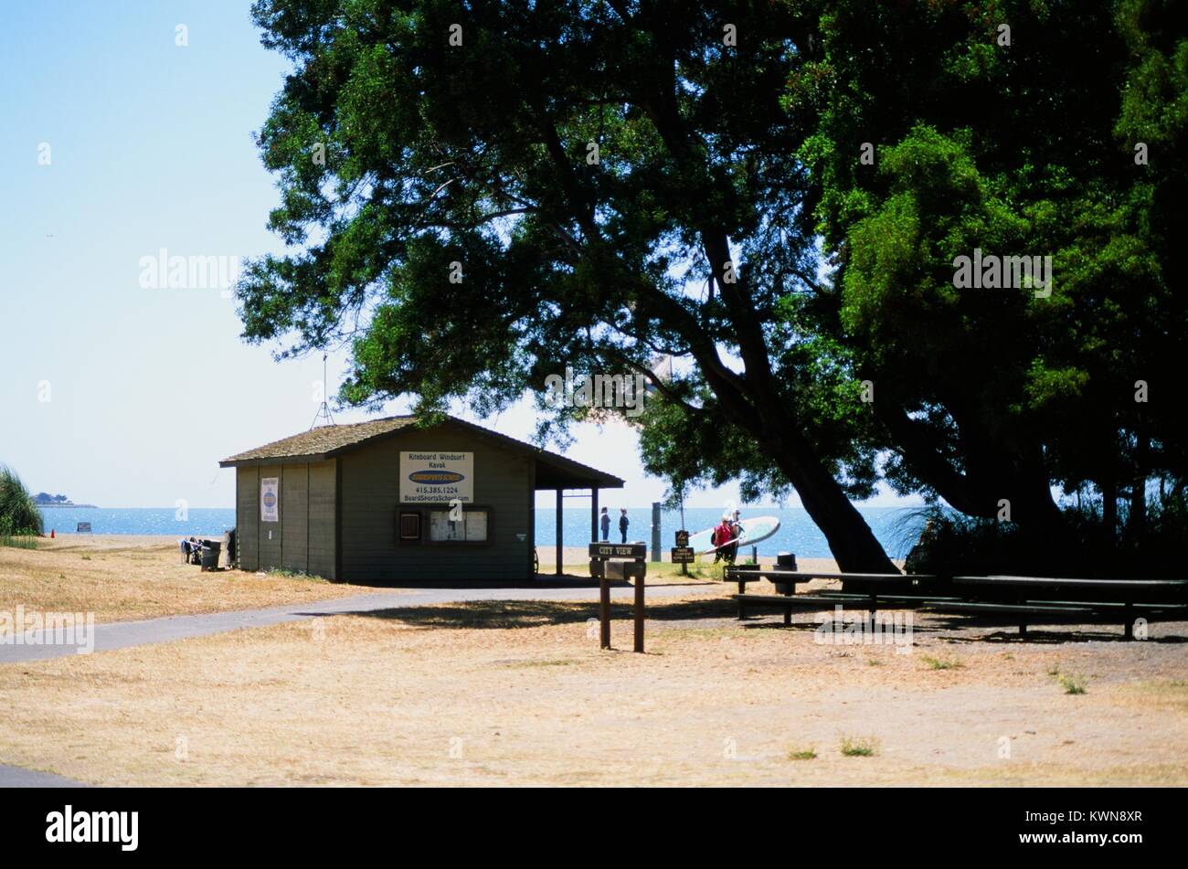 Blick auf die Stadt einen Picknickplatz und Surfbrett/kiteboard Vermietung stand, mit zwei Touristen im Hintergrund hält ein Surfbrett, an der Krone Memorial State Beach, East Bay Regional Park in Alameda, Kalifornien, 20. Juli 2017. Stockfoto