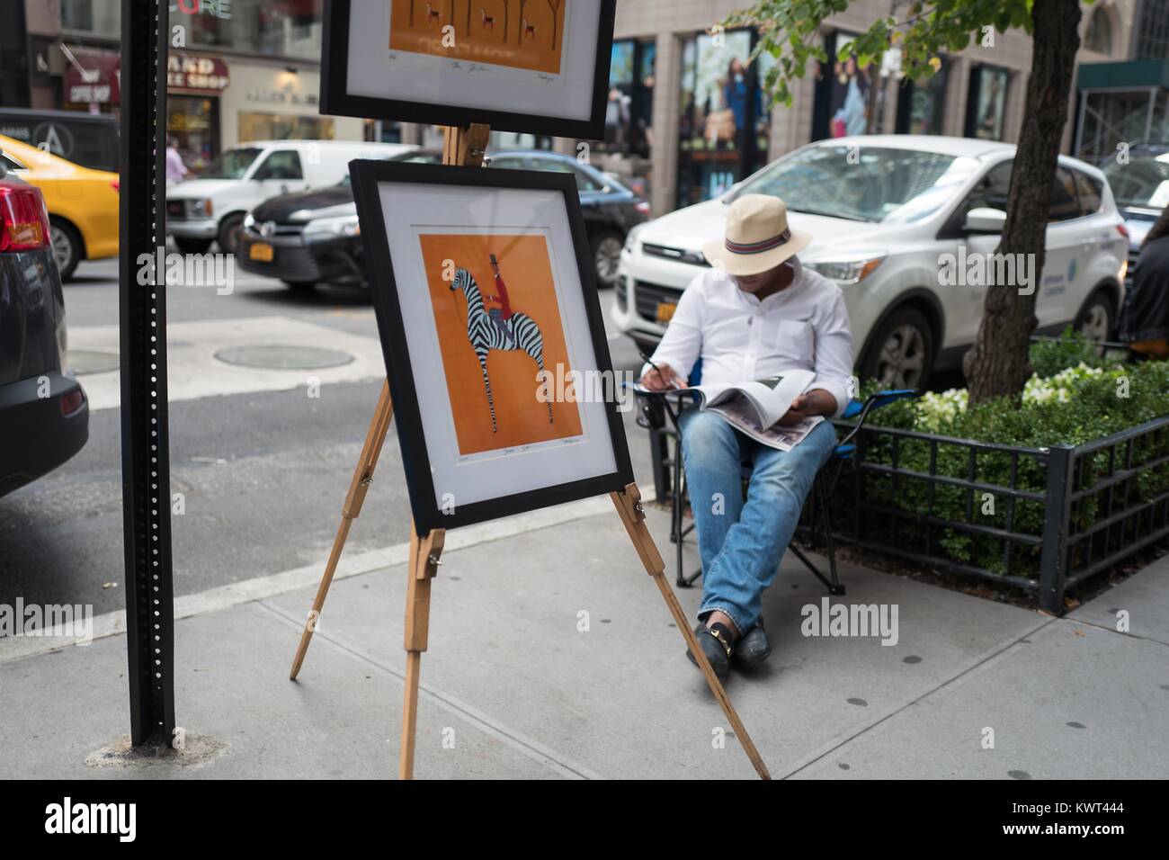 Ein street artist liest eine Zeitschrift, während seine Bilder anzeigen auf der Madison Avenue in der Upper East Side von Manhattan, New York City, New York, 15. September 2017. () Stockfoto