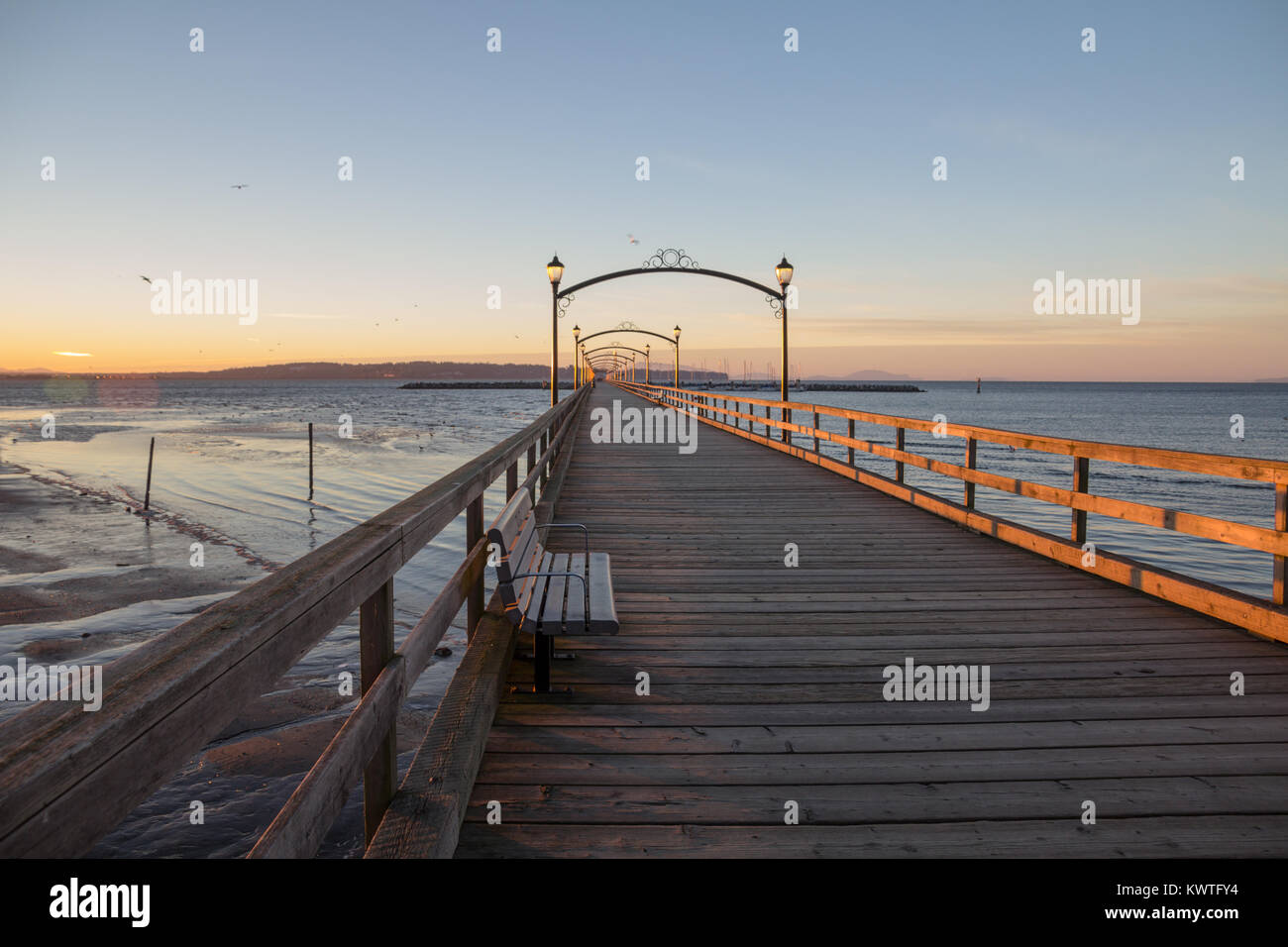 An einem kalten, klaren Wintermorgen, eine Sitzbank ist der Mittelpunkt eines Fotos von Pier's White Rock. Die hellen Palette eines Winters sunrise bringt Farbe. Stockfoto