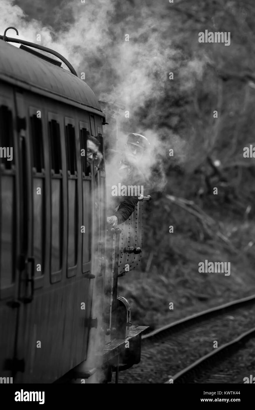 Schwarz & Weiß Portraitfotos von sich bewegenden Dampfzug auf Erbe Bahnstrecke. Der Fahrer steht auf Fußplatte, lehnte sich aus Loco, verdeckt von Dampf. Stockfoto