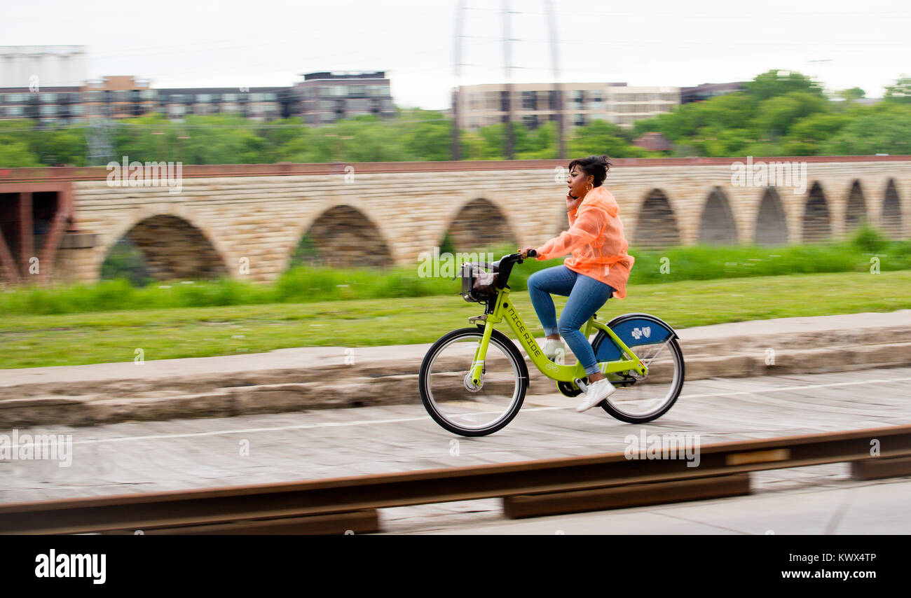 Minneapolis, Minnesota/USA - 05/24/17 Dokumentarfilm Editorial. Afrikanische amerikanische Frau auf Handy beim Reiten Fahrradverleih Fahrrad; Steinbogenbrücke. Stockfoto