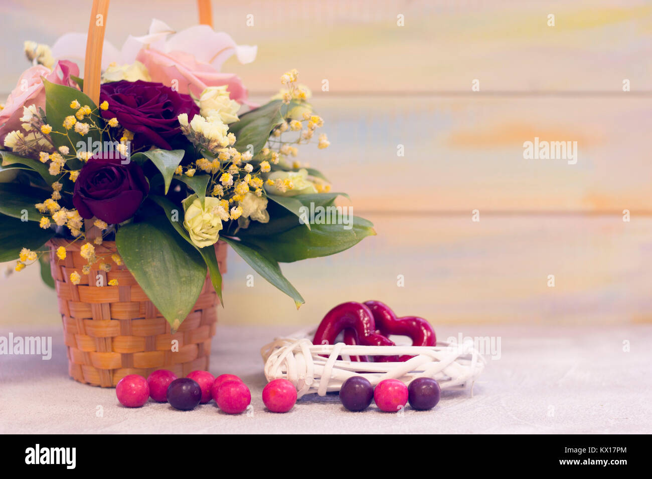 Romantische Stillleben mit einem Korb von duftenden Rosen, Rattan Kranz mit roten Herzen innerhalb und farbigen runde Bonbons auf dem Tisch. Helle Holz- backgrou Stockfoto