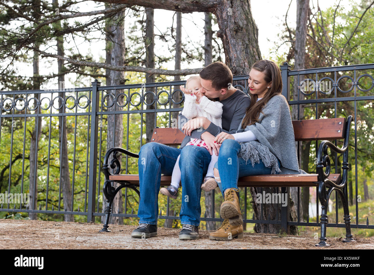 Glückliche Familie von drei sitzen auf einer Bank in einem Park ihre Zeit zusammen genießen. Vater einen Kuss zu seinem Kind Tochter, Mutter lächelnd. Stockfoto